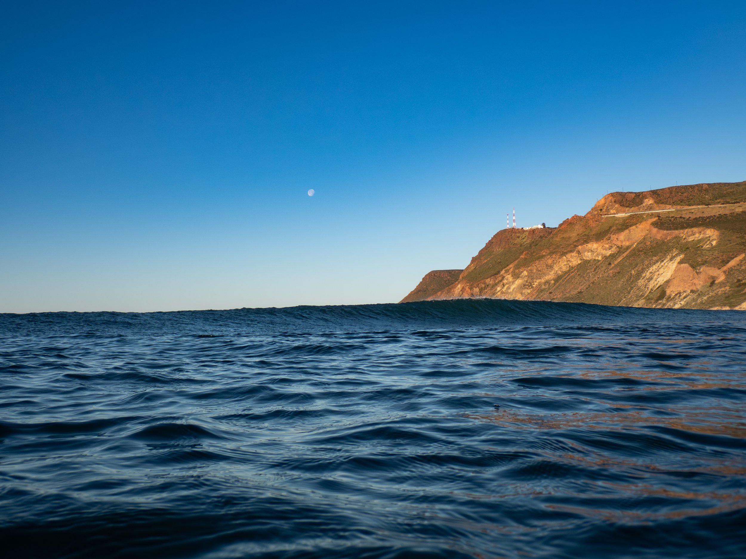 Ocean waves near a coastal hillside under a clear blue sky with the moon visible.