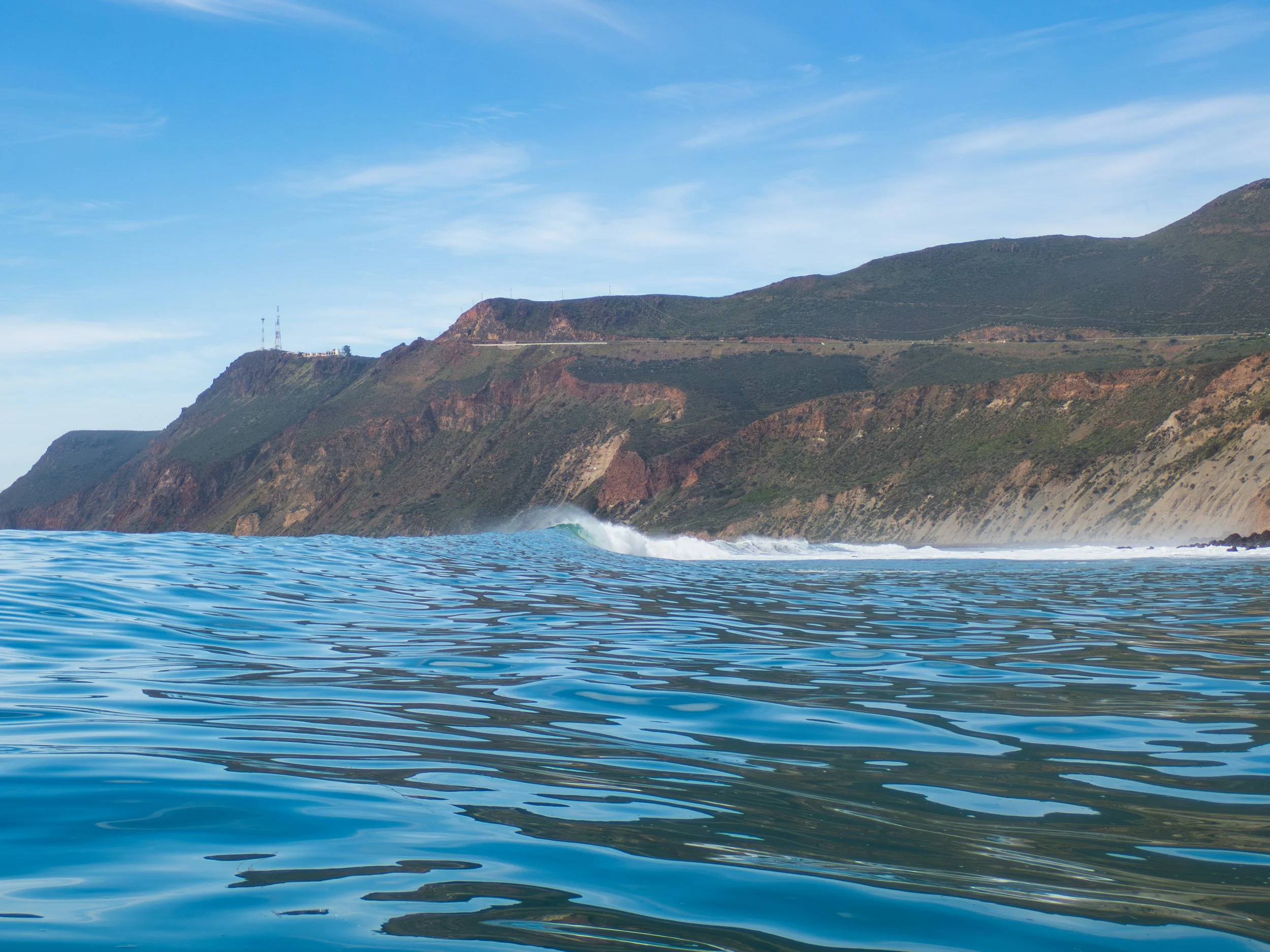 Ocean water with small waves near rolling coastal cliffs under a partly cloudy sky.