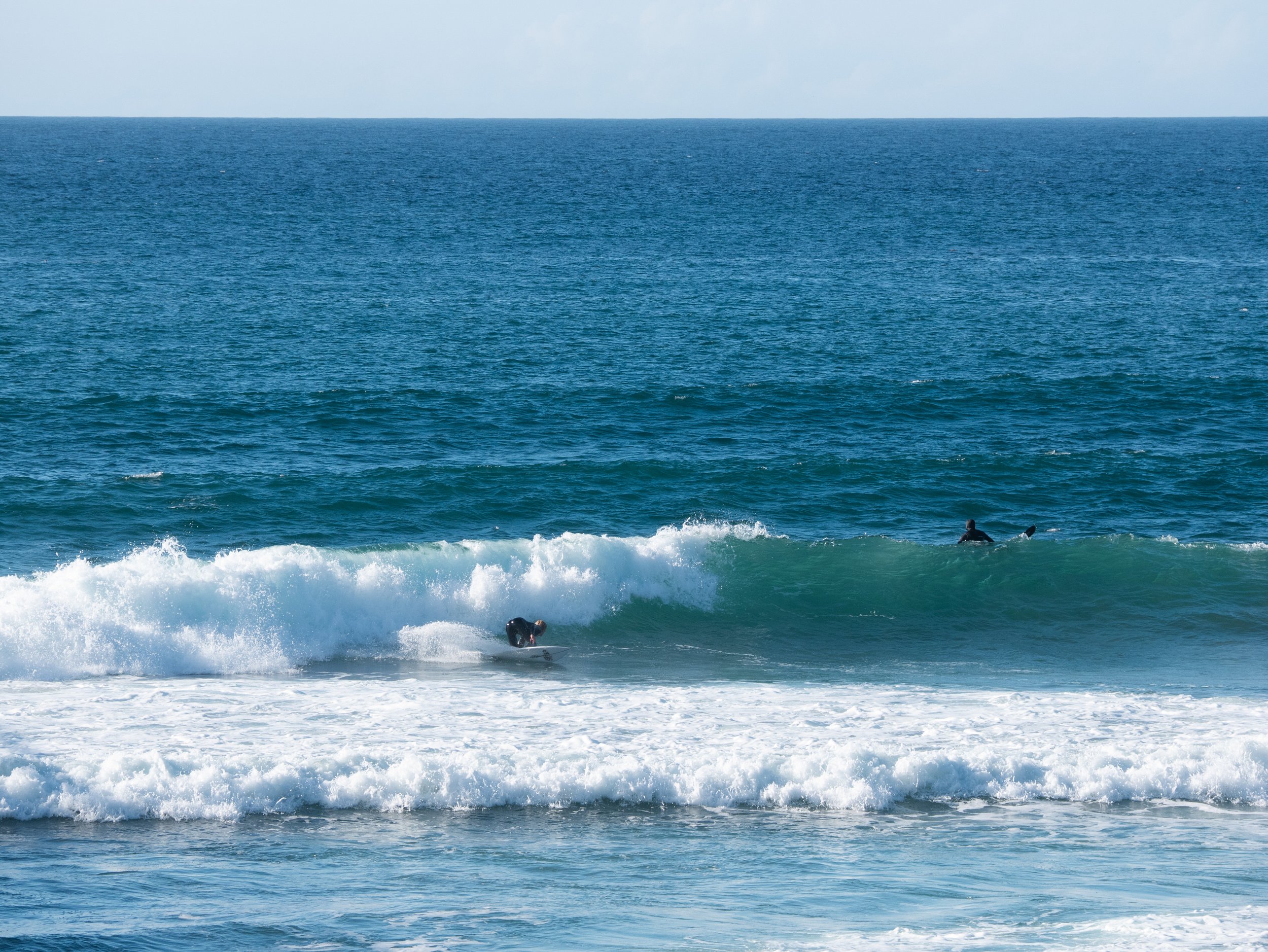 Two surfers in wetsuits riding waves on the ocean, with a vast blue sea and sky in the background.