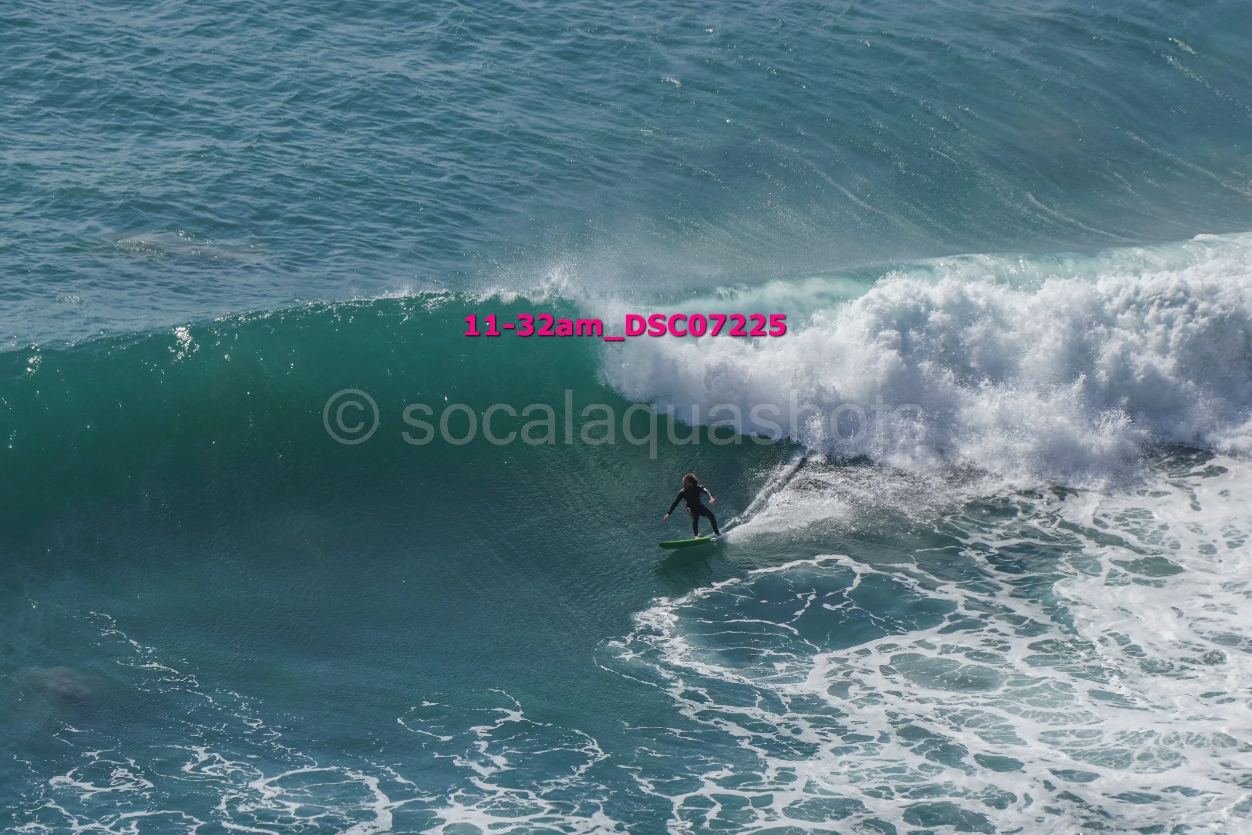 A person surfing on a large ocean wave with white foam, wearing a black wetsuit, on a green surfboard.
