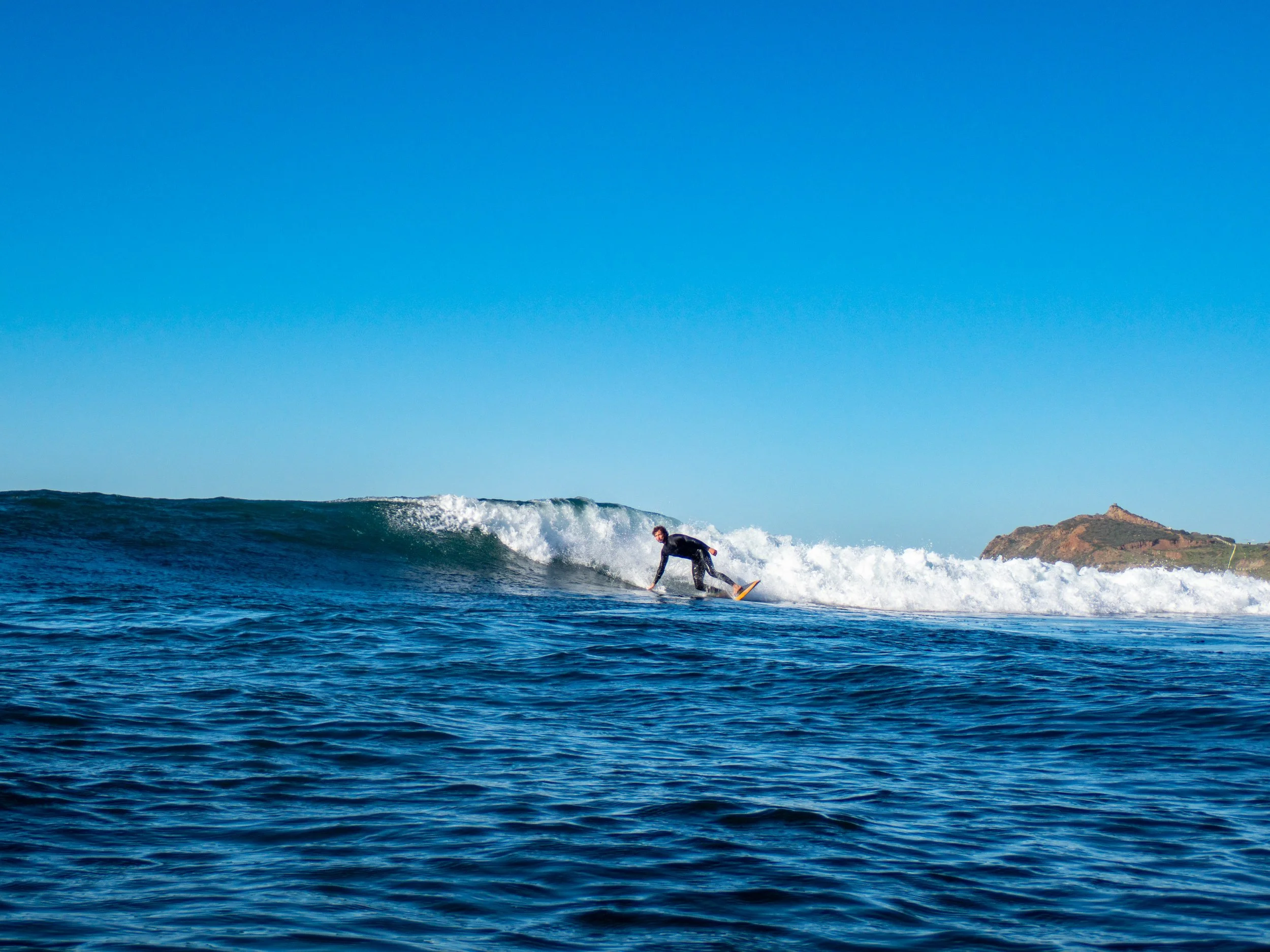 A person surfing on a wave in the ocean with a hilly landscape in the background under a clear blue sky.