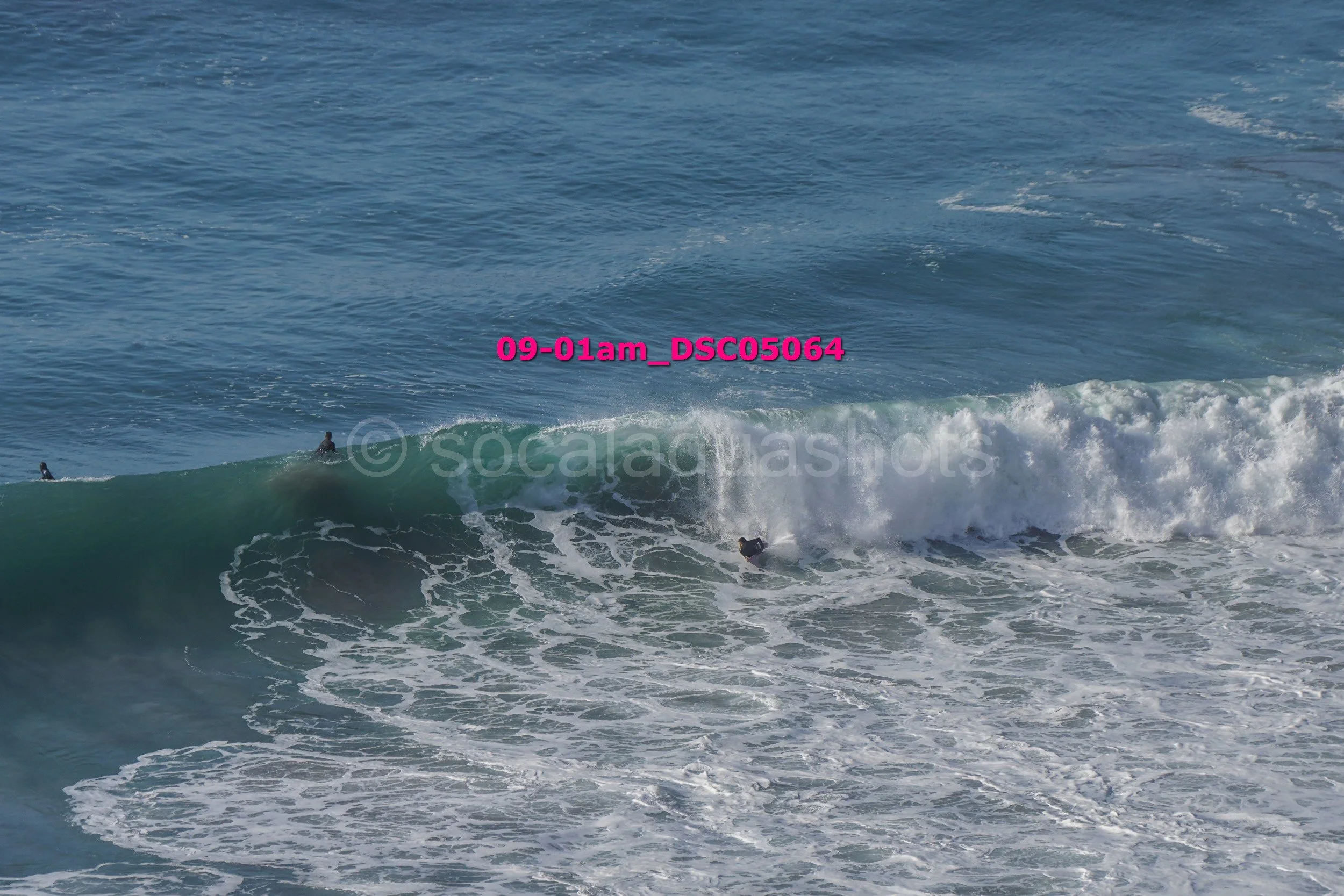 Surfers riding and drifting on ocean waves with white foam and ripples, blue water, and a clear sky.