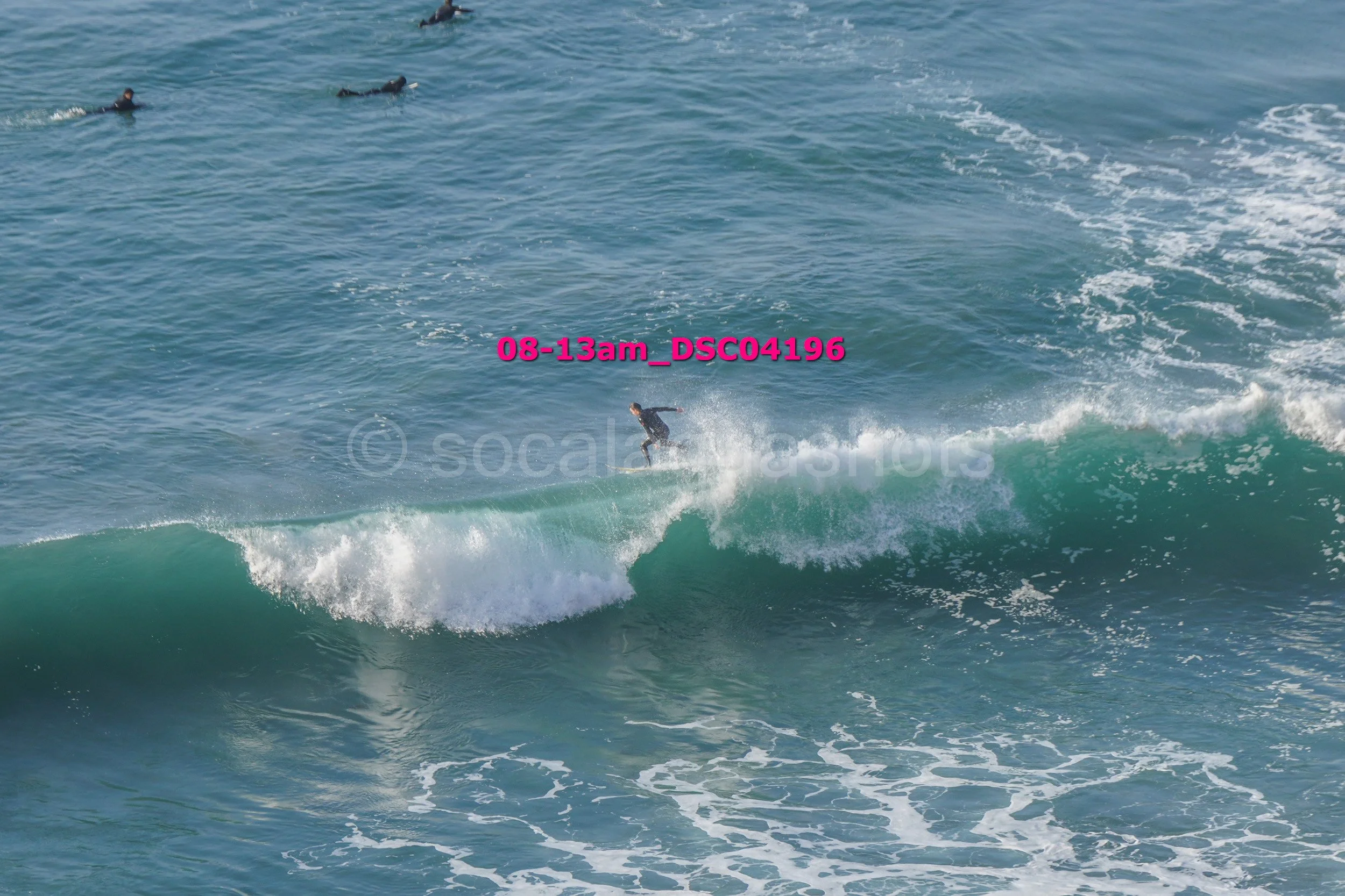 A person surfing on a large wave in the ocean with two other surfers in the background and pink text overlay reading '08-13am_DSC04196'.