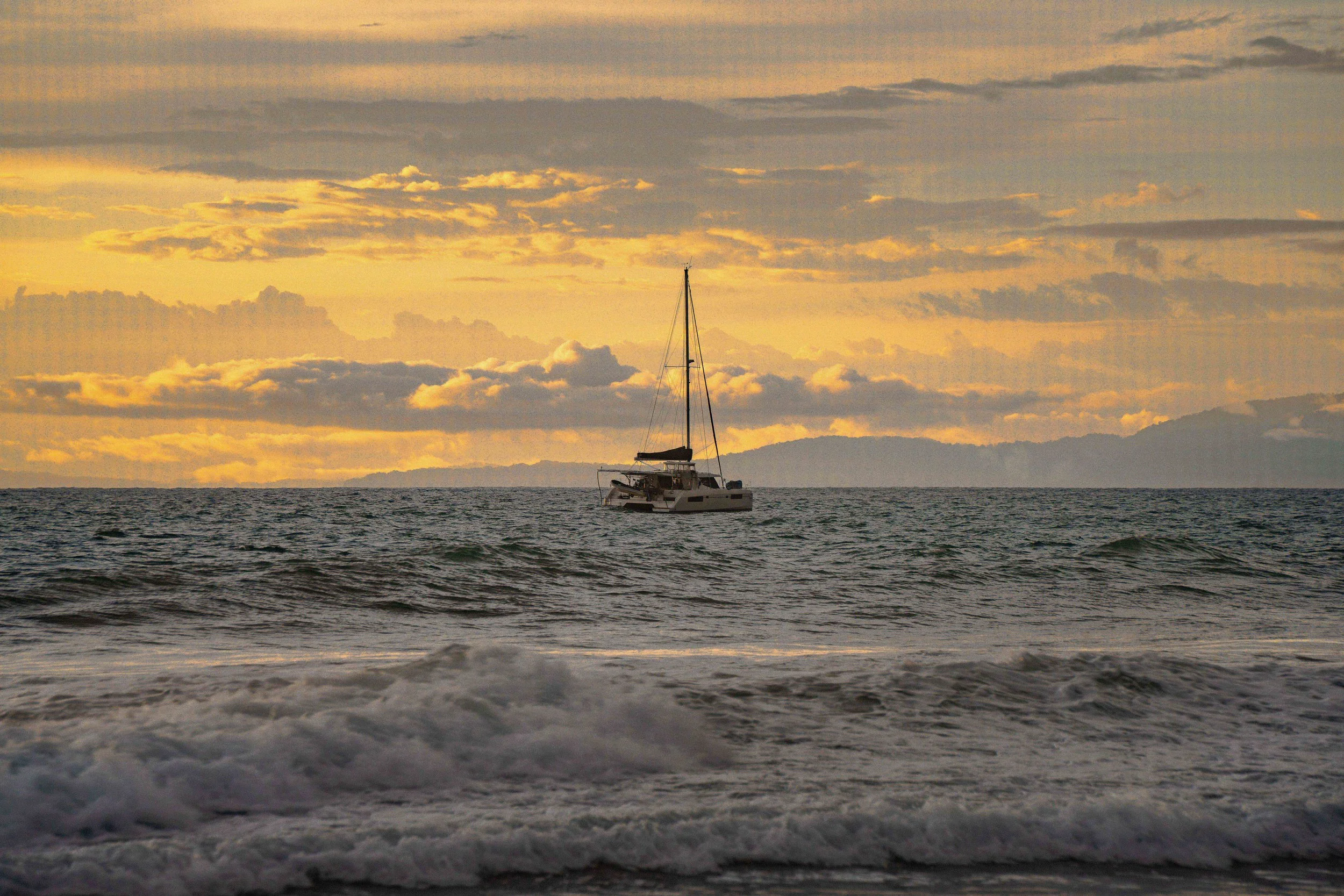 A sailboat floating on the ocean at sunset with clouds in the sky and mountains in the background.