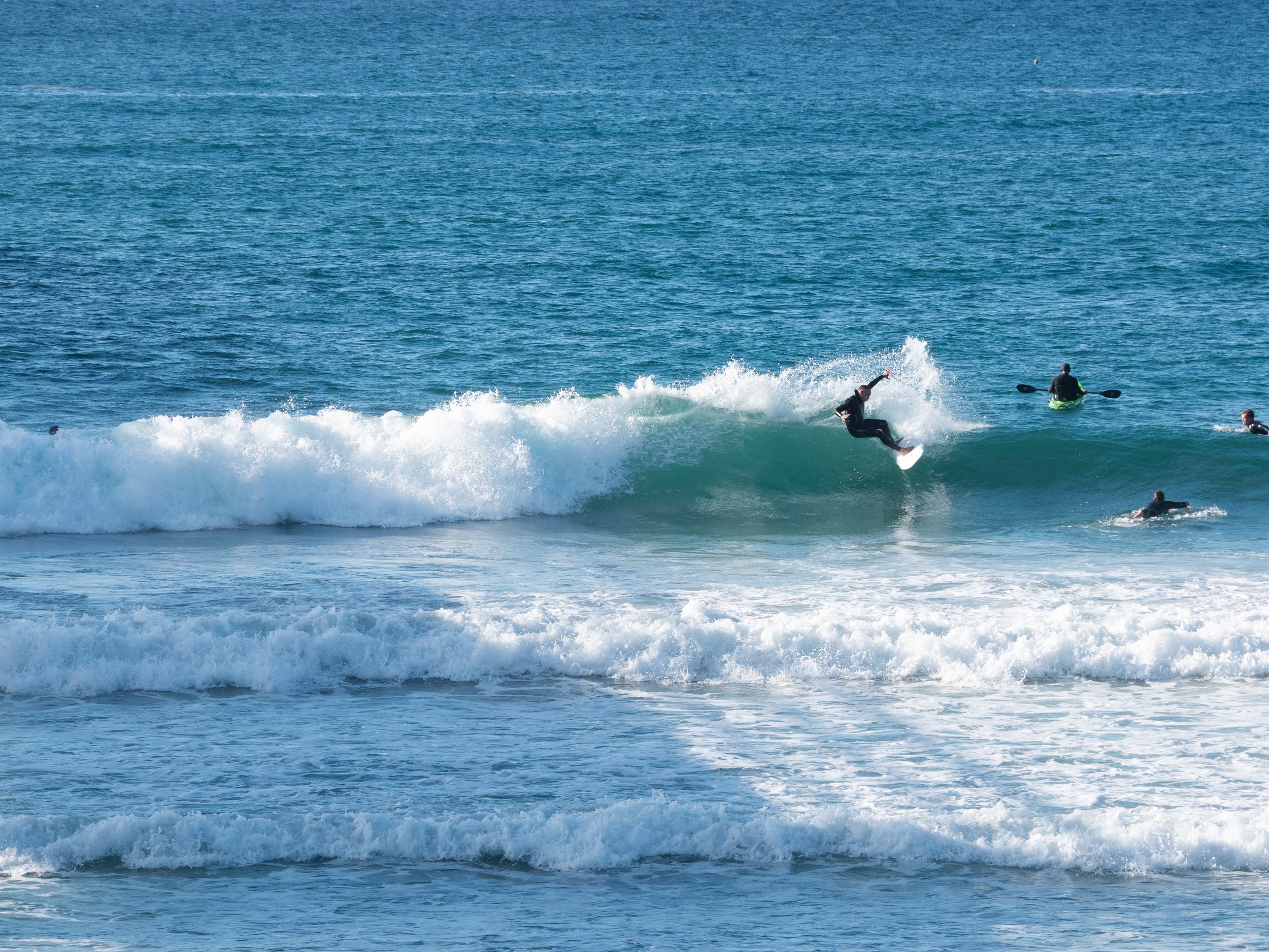 Surfer riding a wave at the beach with several people kayaking or paddleboarding nearby.