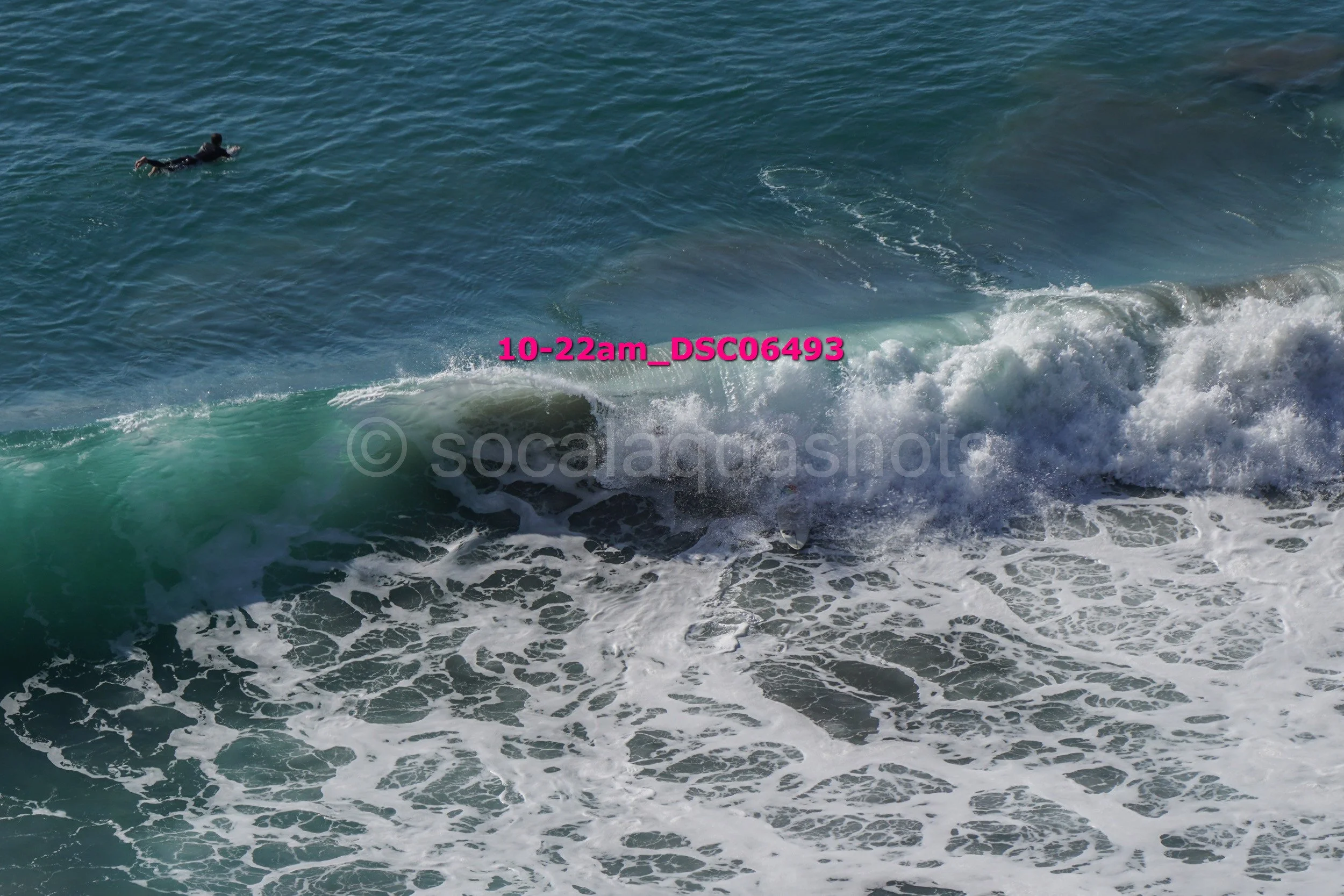 A person in a wetsuit swimming in the ocean near a breaking wave, with a camera watermark and the text '10-22am_DSC6493' overlaid on the image.