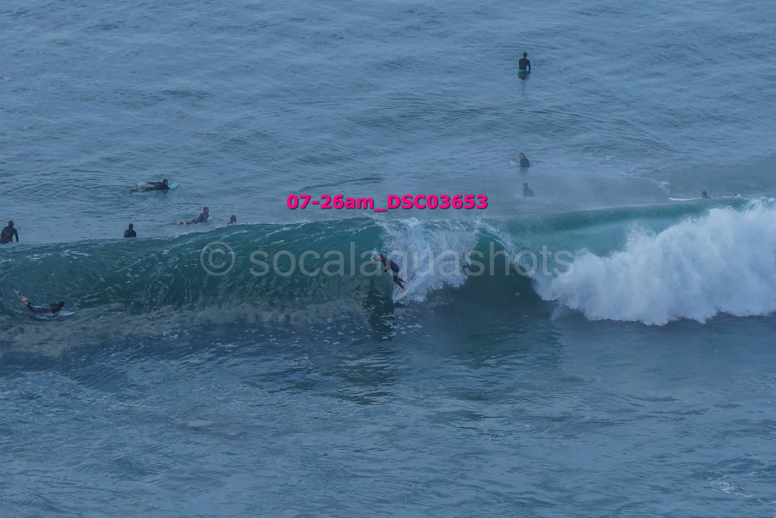 A surfer riding a wave with multiple people in the water around him, some on surfboards and others standing in the ocean.