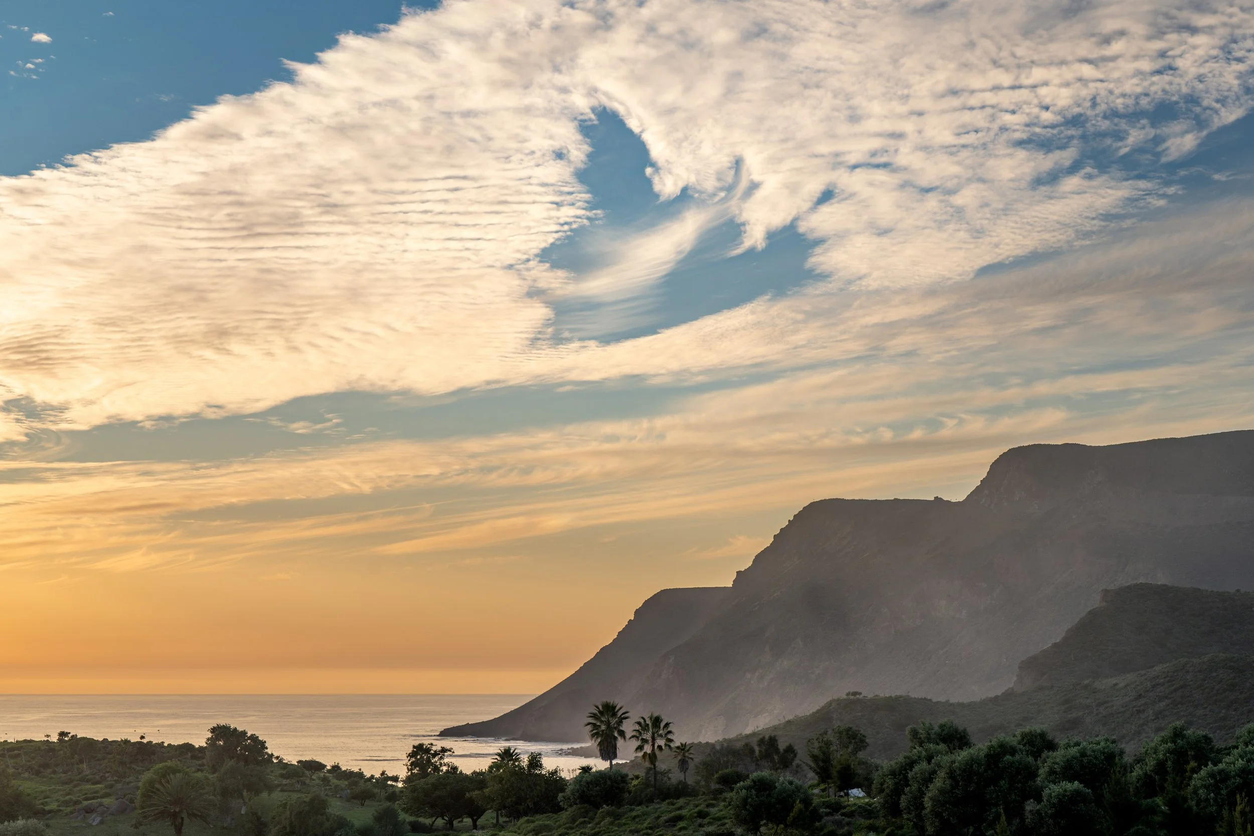 Sunset over a coastal landscape with mountains, trees, and a partly cloudy sky.