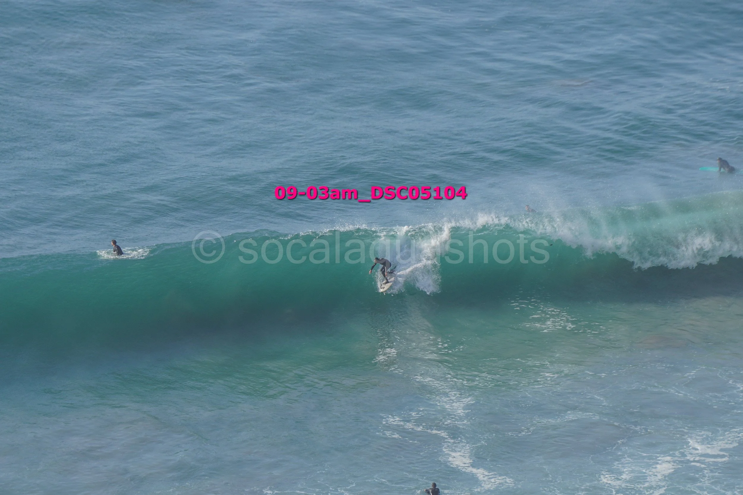 A person surfing on a large ocean wave with two other surfers nearby in the water.