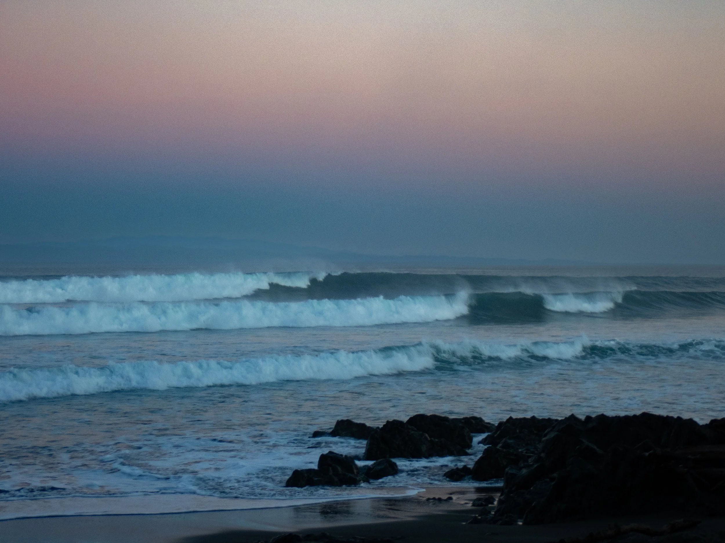 Ocean waves crashing against rocks at sunset with colorful sky in the background.