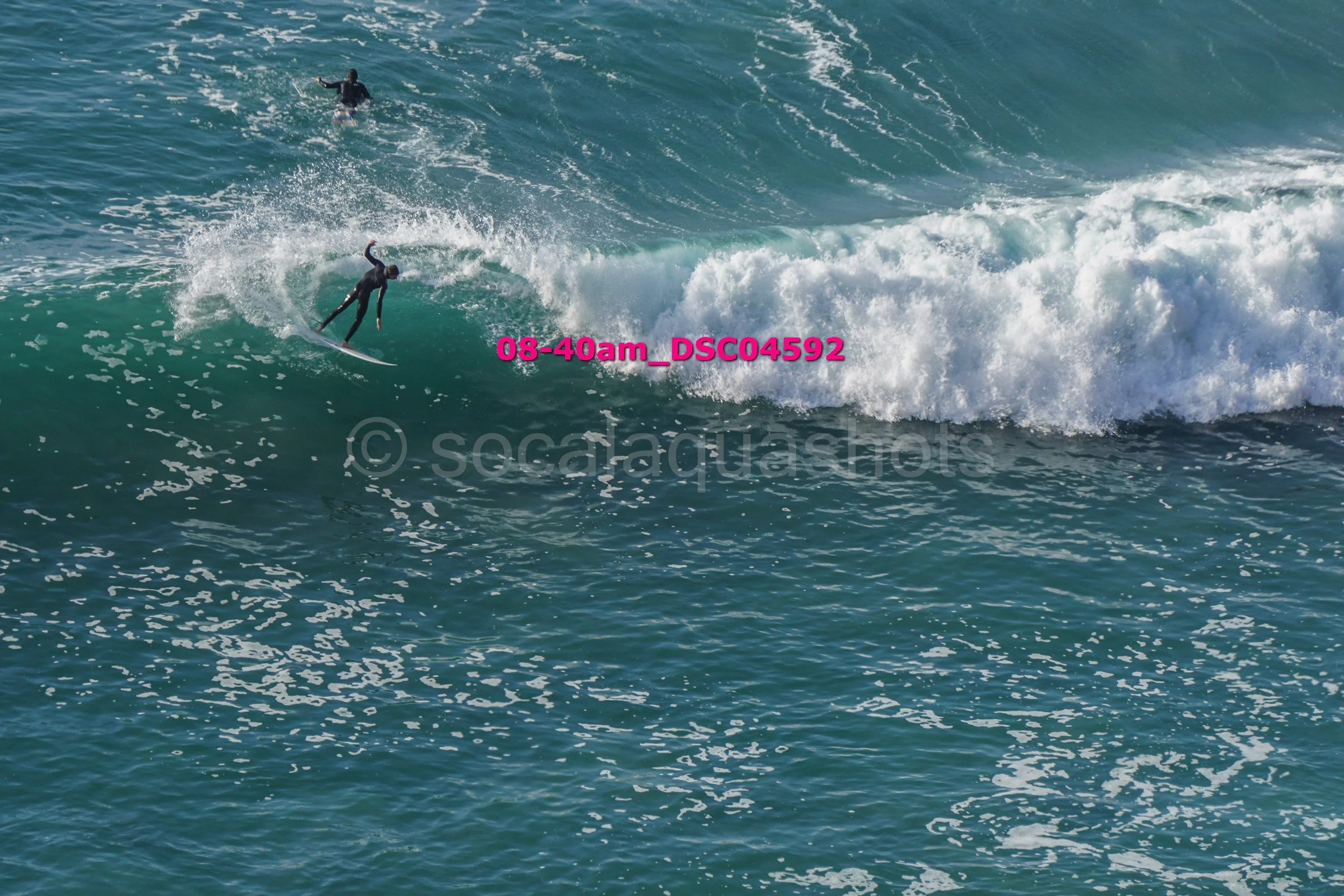 A person in a wetsuit surfing on a wave in the ocean, with another surfer in the background.