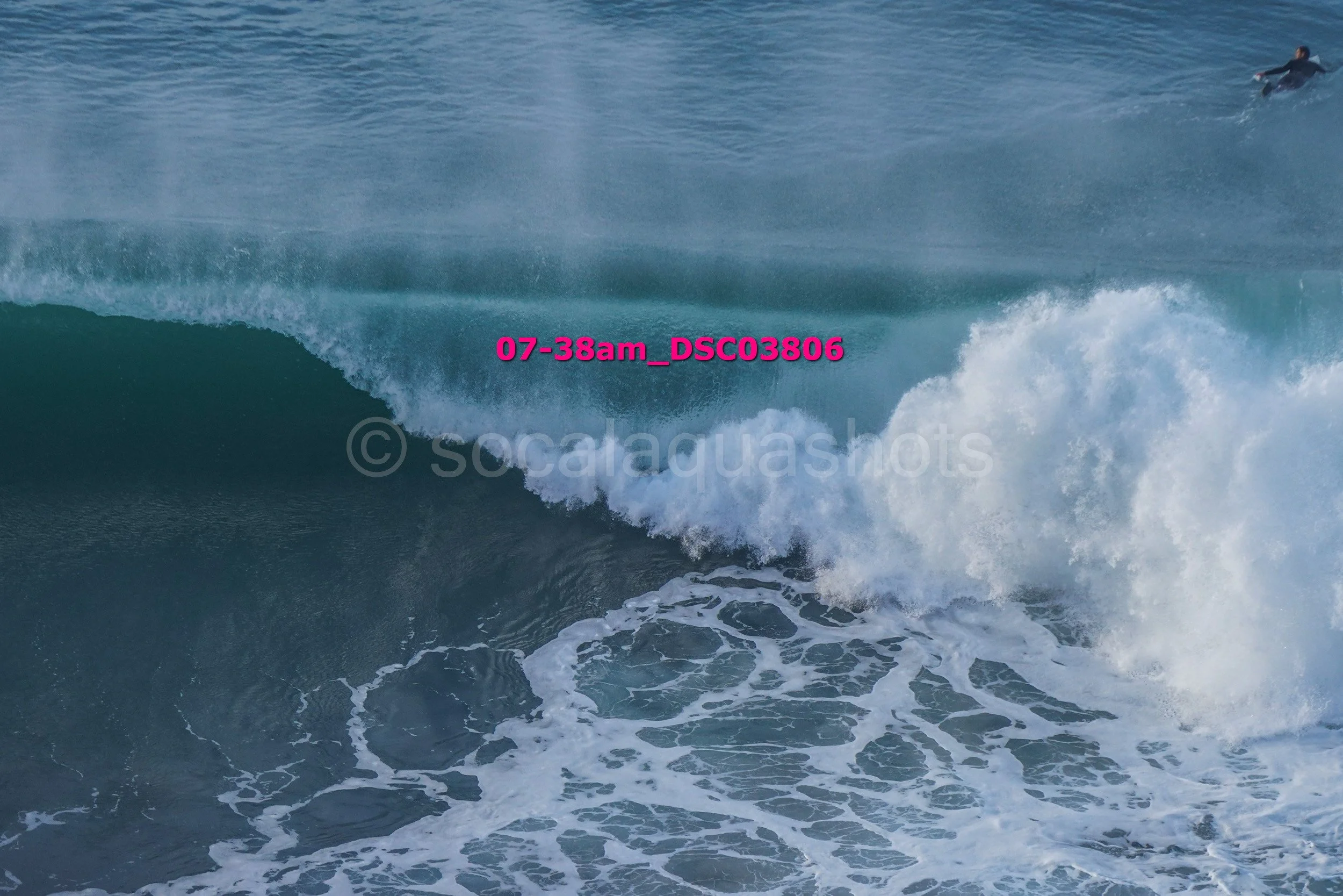 Large ocean wave breaking with a surfer in the background and a wave crest in the foreground.