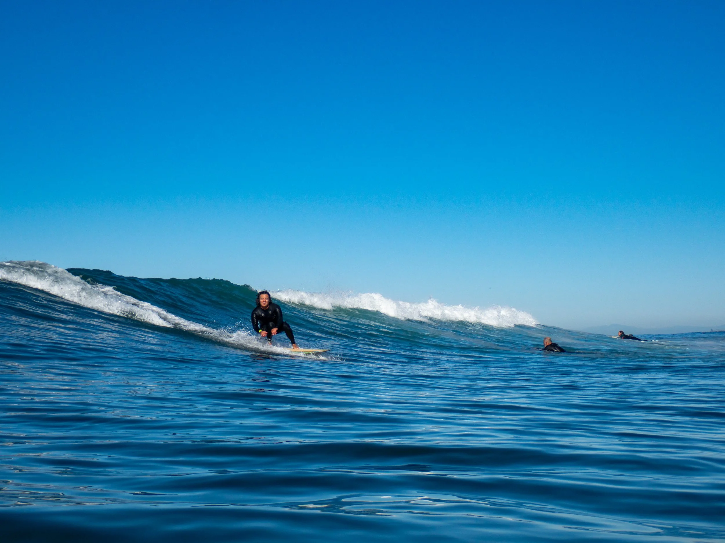 A woman surfing on a wave in the ocean, with two other surfers visible in the distance and a clear blue sky above.