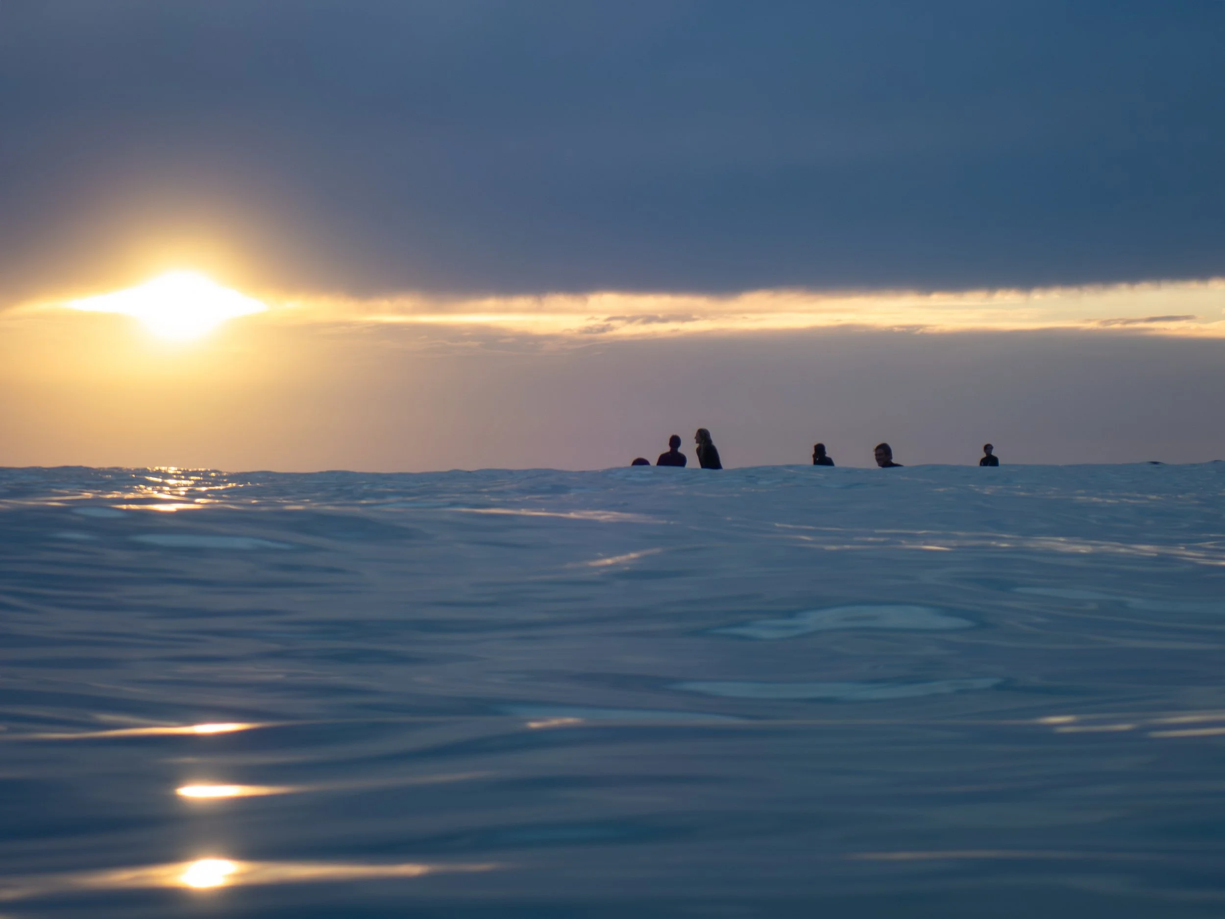 People swimming or floating in the ocean during sunset, with a sky partly covered by dark clouds.