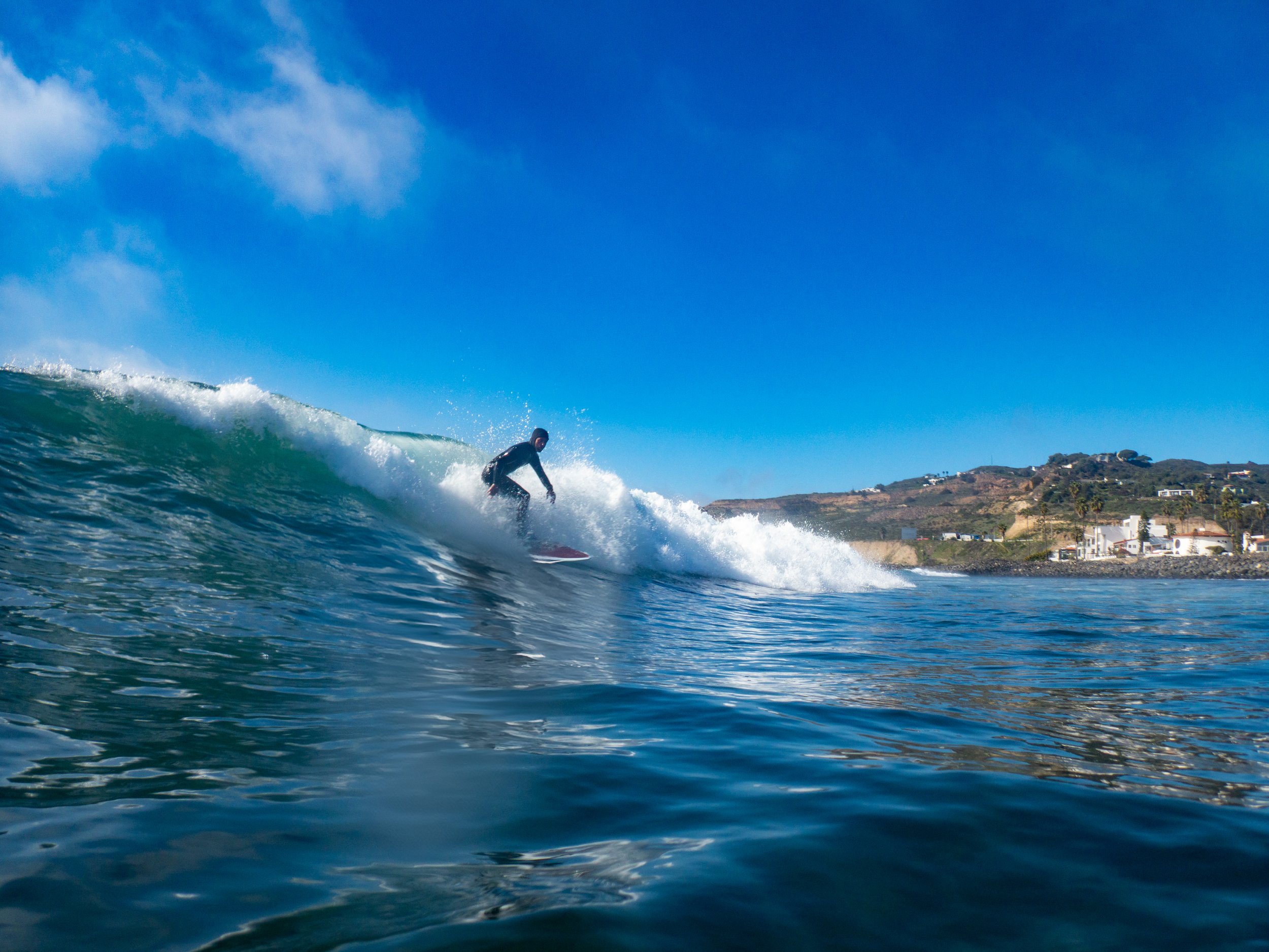 A person surfing on a wave in the ocean with a coastline and houses in the background on a sunny day.