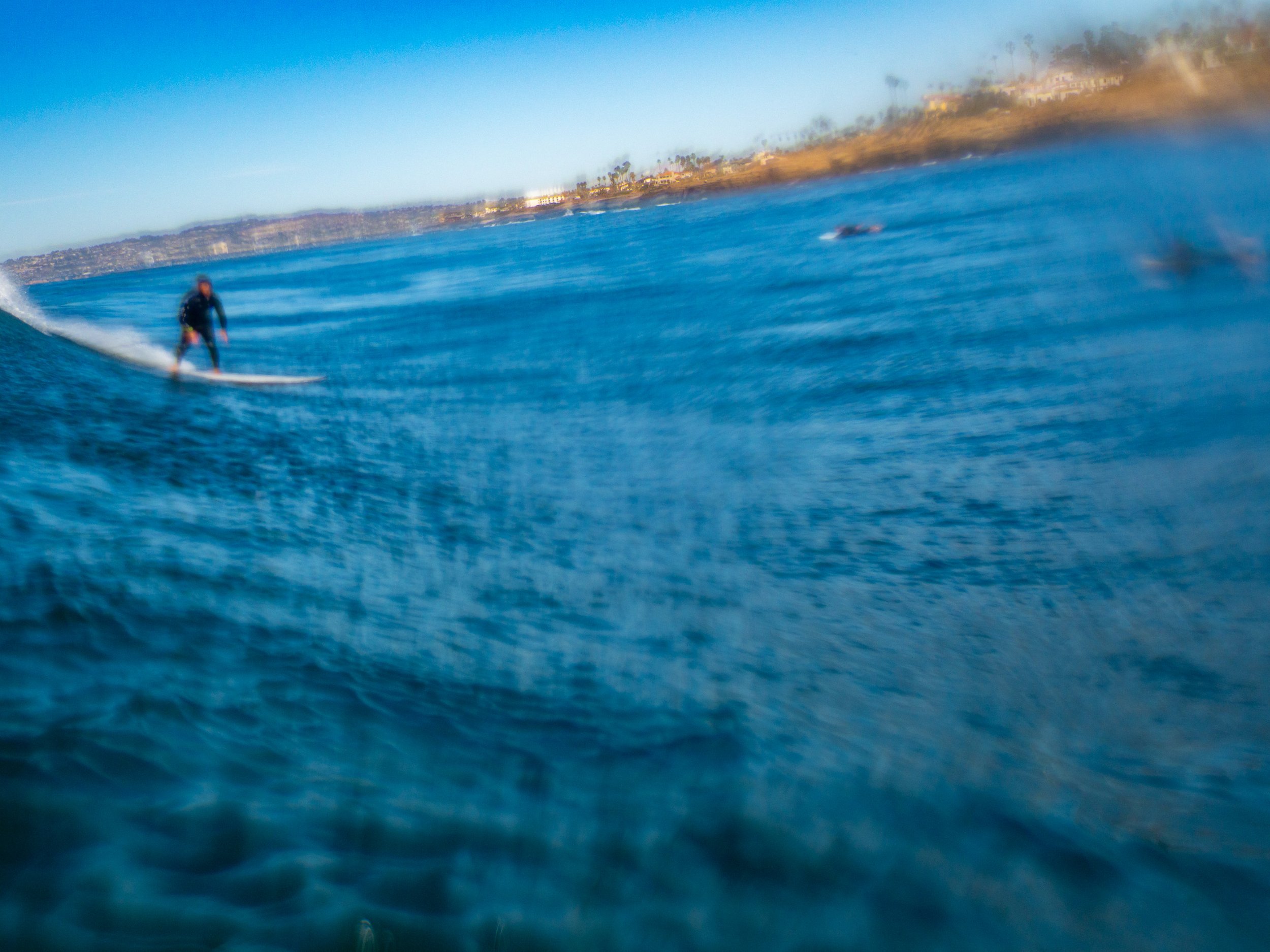 A person riding a wave on a paddleboard in the ocean, with a distant shoreline and clear blue sky.