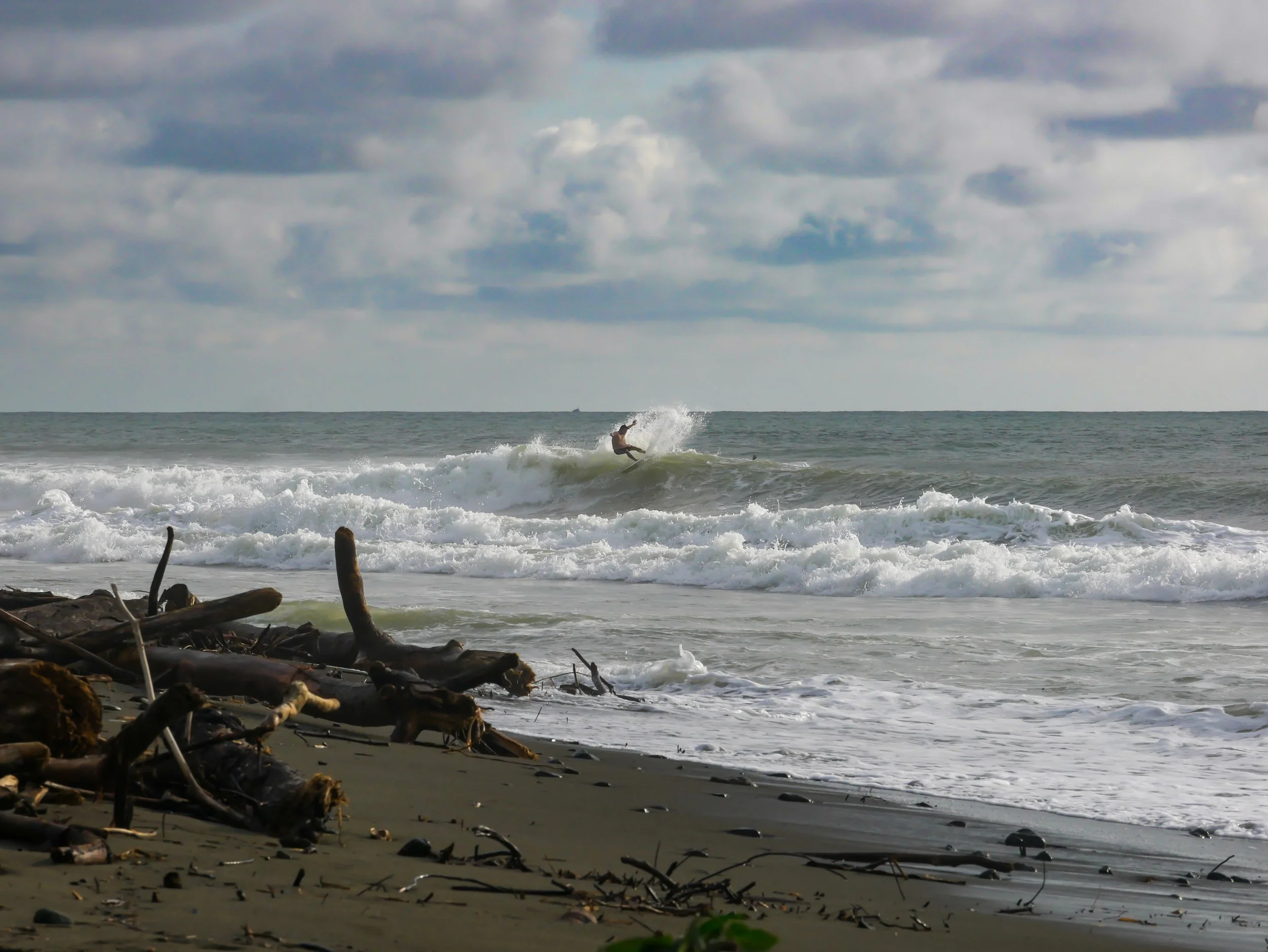 Surfer riding waves near shore with driftwood on beach and cloudy sky.