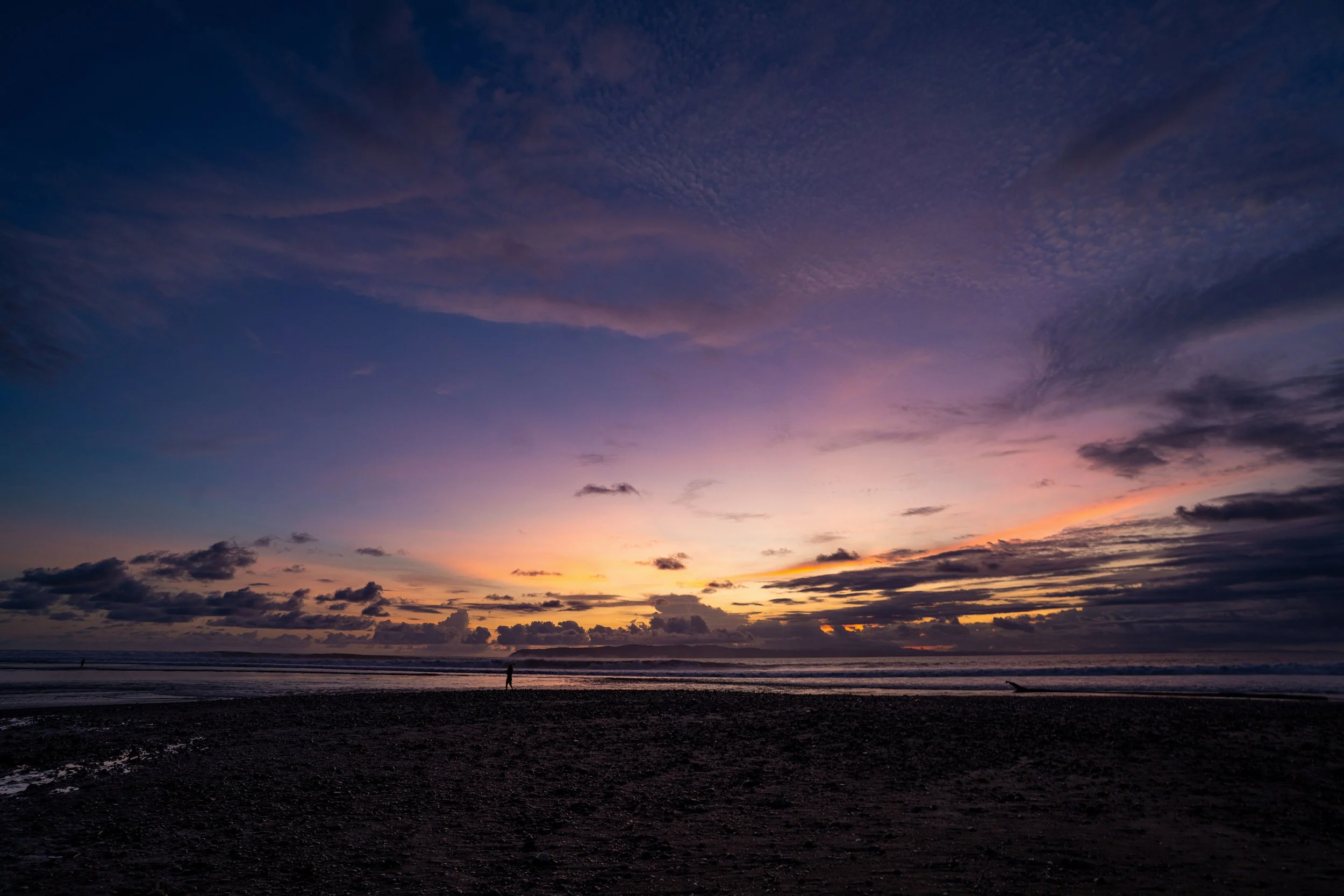 A beach at sunset with colorful sky, clouds, and a person walking along the shoreline.