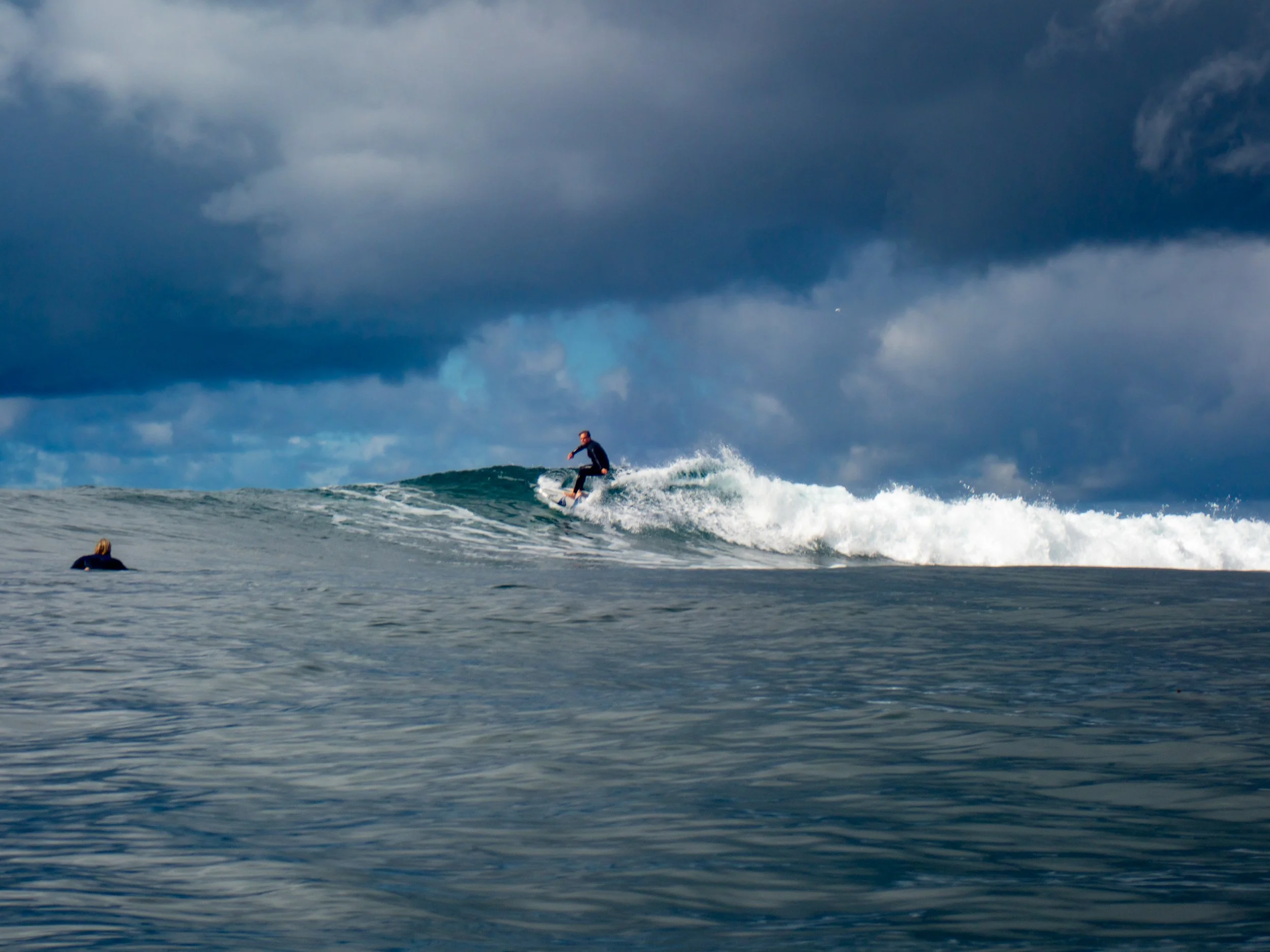 A person surfing on a large wave in the ocean with dark clouds overhead.