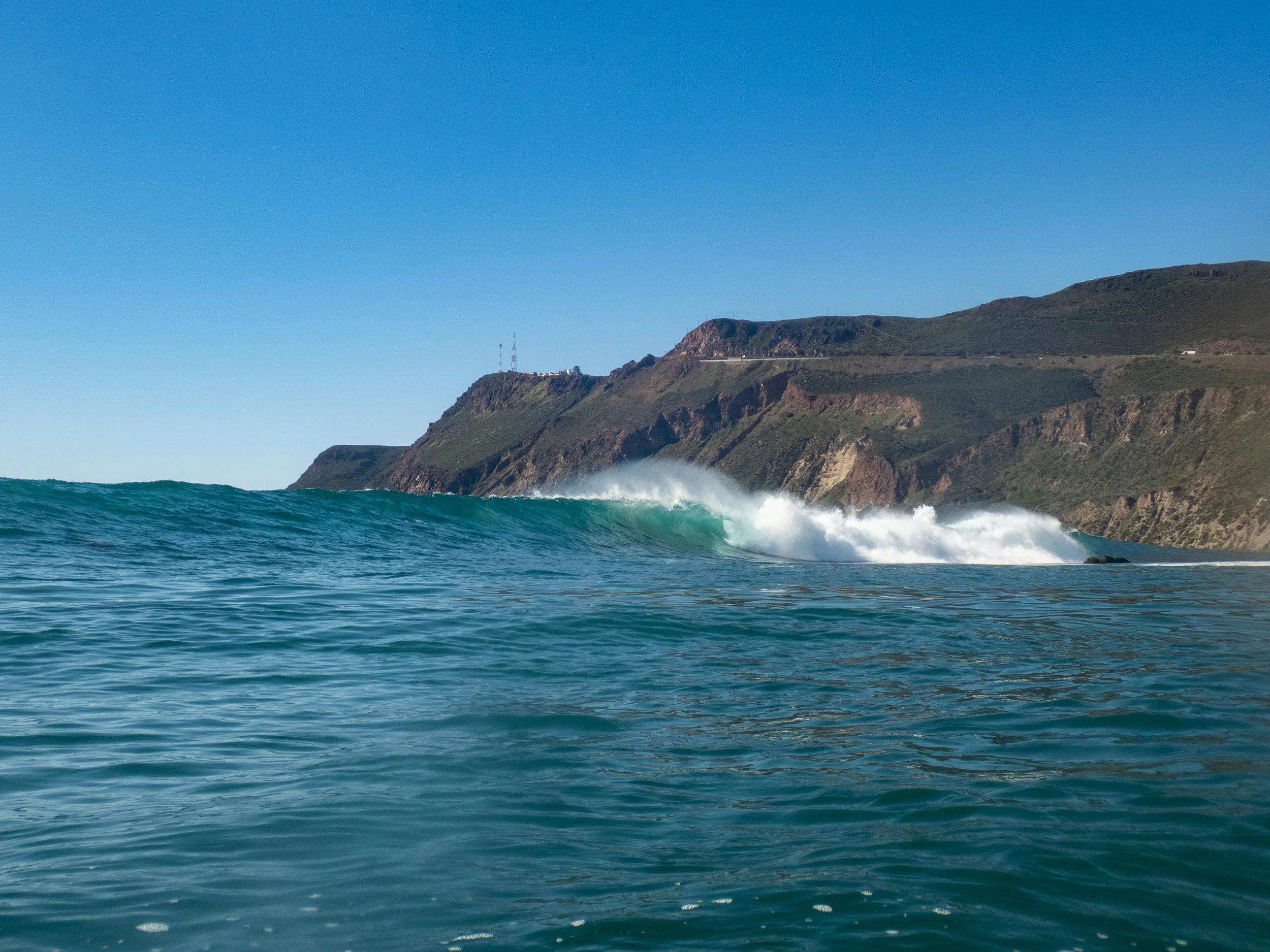 Ocean waves near a rugged coastline with hills and a clear blue sky.