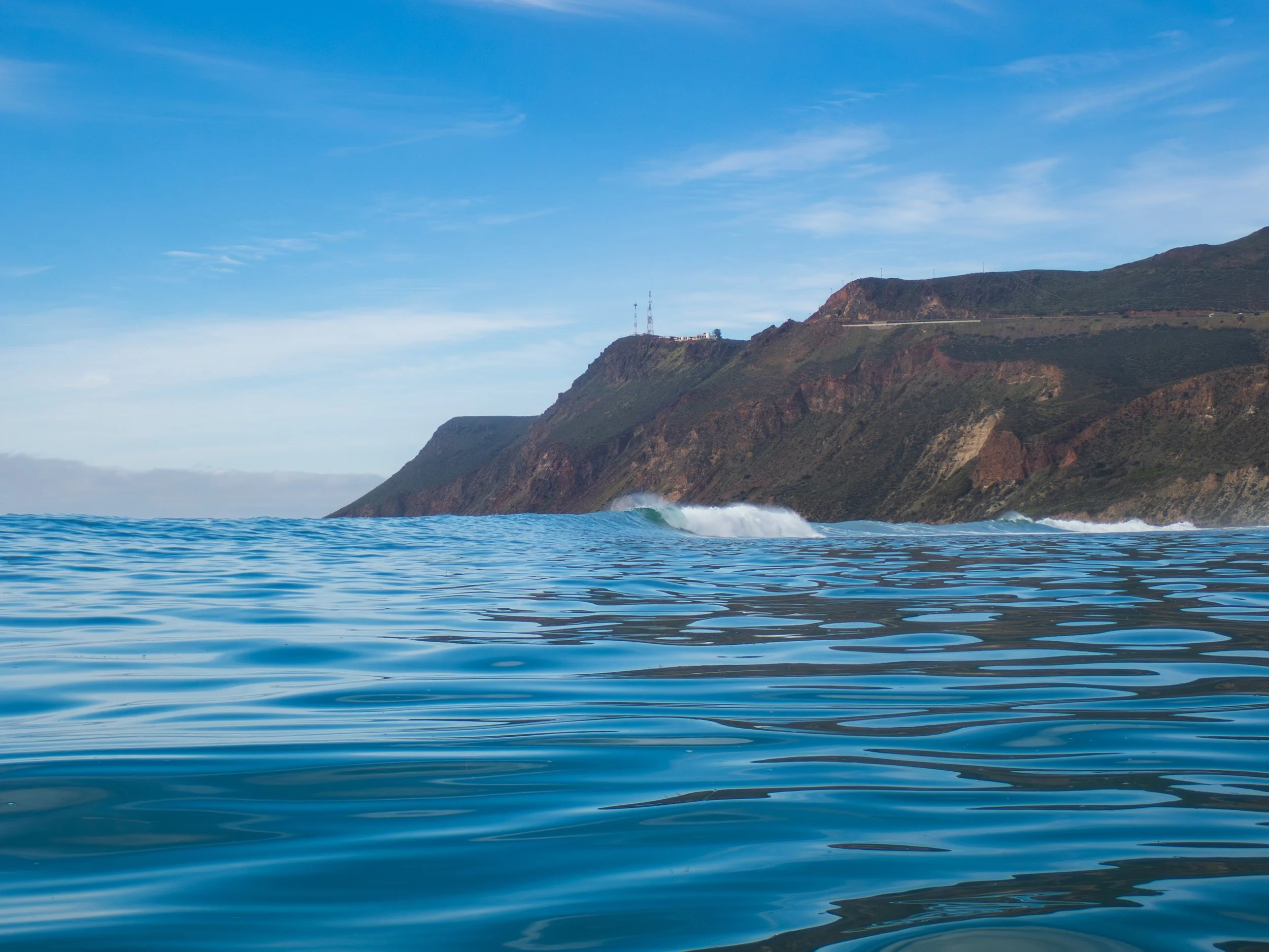 View of the ocean with gentle waves and a coastline with cliffs and hills under a partly cloudy sky.