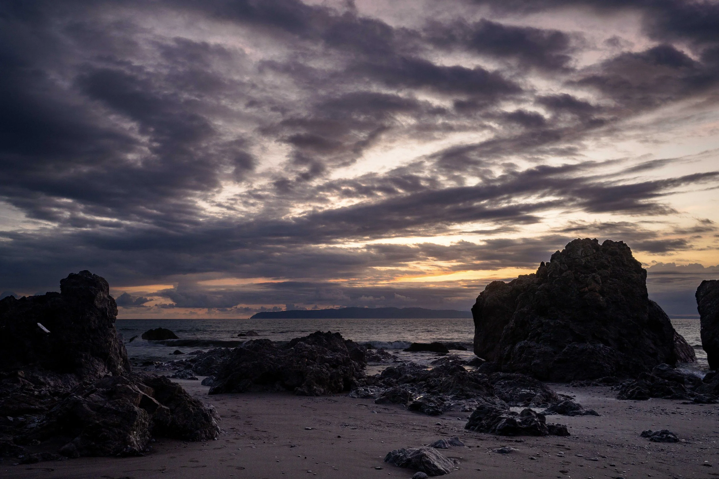 Sunset over the ocean with cloudy sky and large rocks on a sandy beach in the foreground