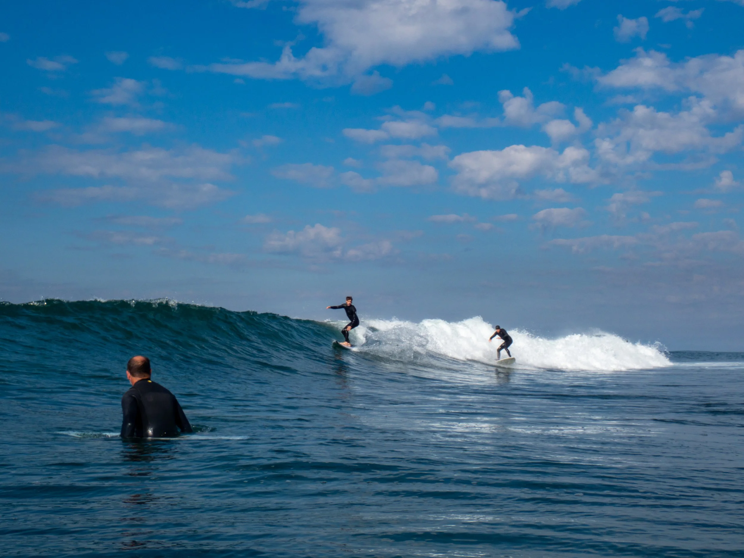 Two surfers riding a wave in the ocean while a man in a wetsuit watches from the water.
