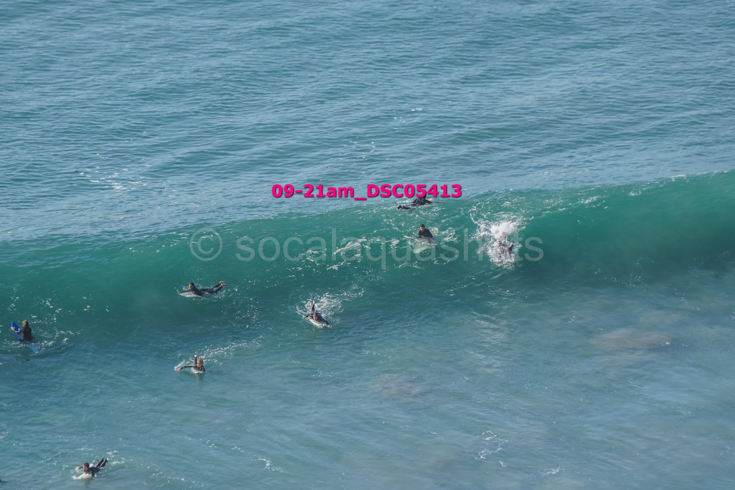 Surfers in wetsuits riding and swimming alongside large ocean waves with a blue sky background.