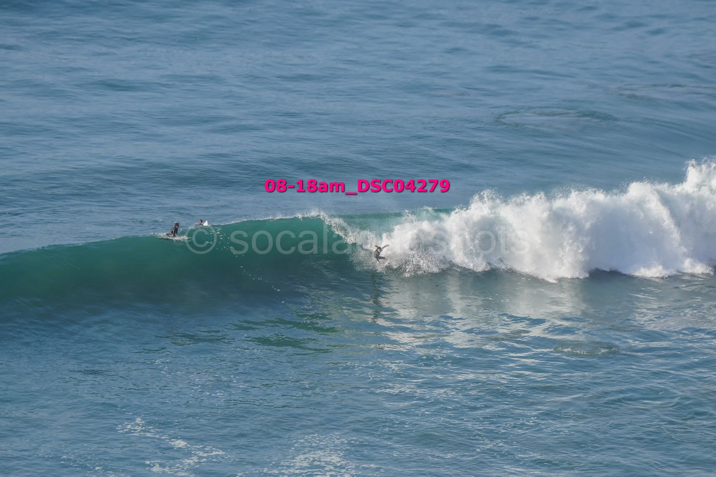 Two surfers riding a large wave in the ocean, with one surfer on the wave and another waiting on a surfboard in the water, under a clear sky.