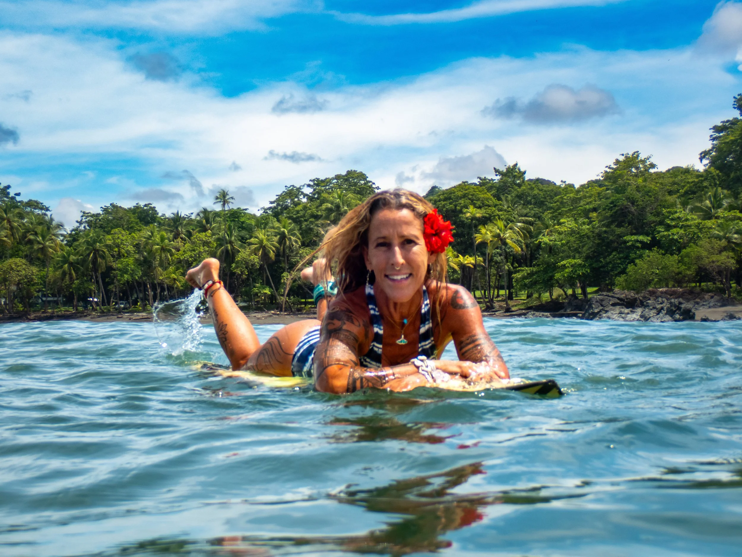 Woman lying on a surfboard in the water with tropical trees and blue sky in the background, wearing a striped swimsuit and a red flower in her hair.