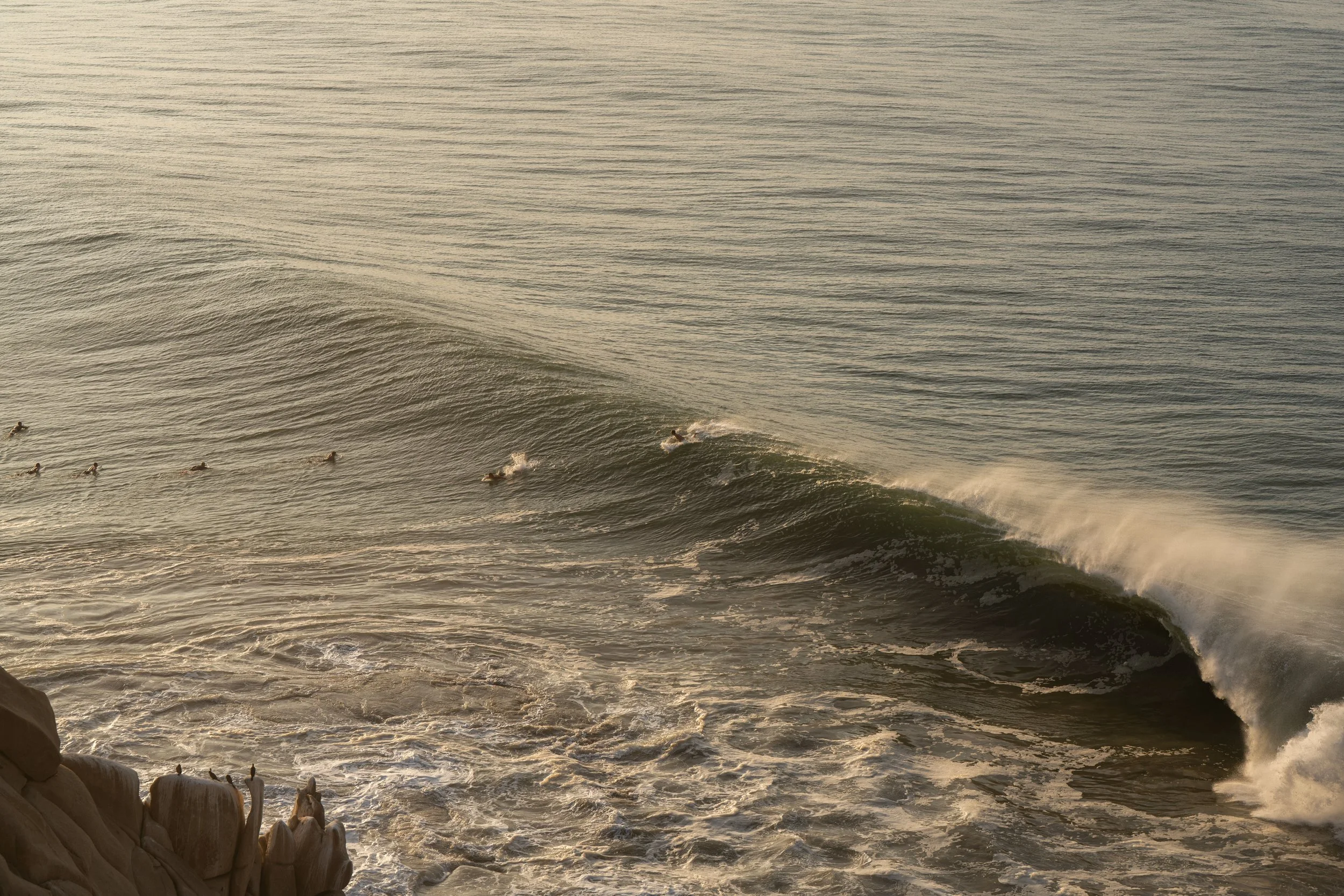 A view of the ocean with gentle waves and a few surfers riding the waves near the shore, captured at sunset.