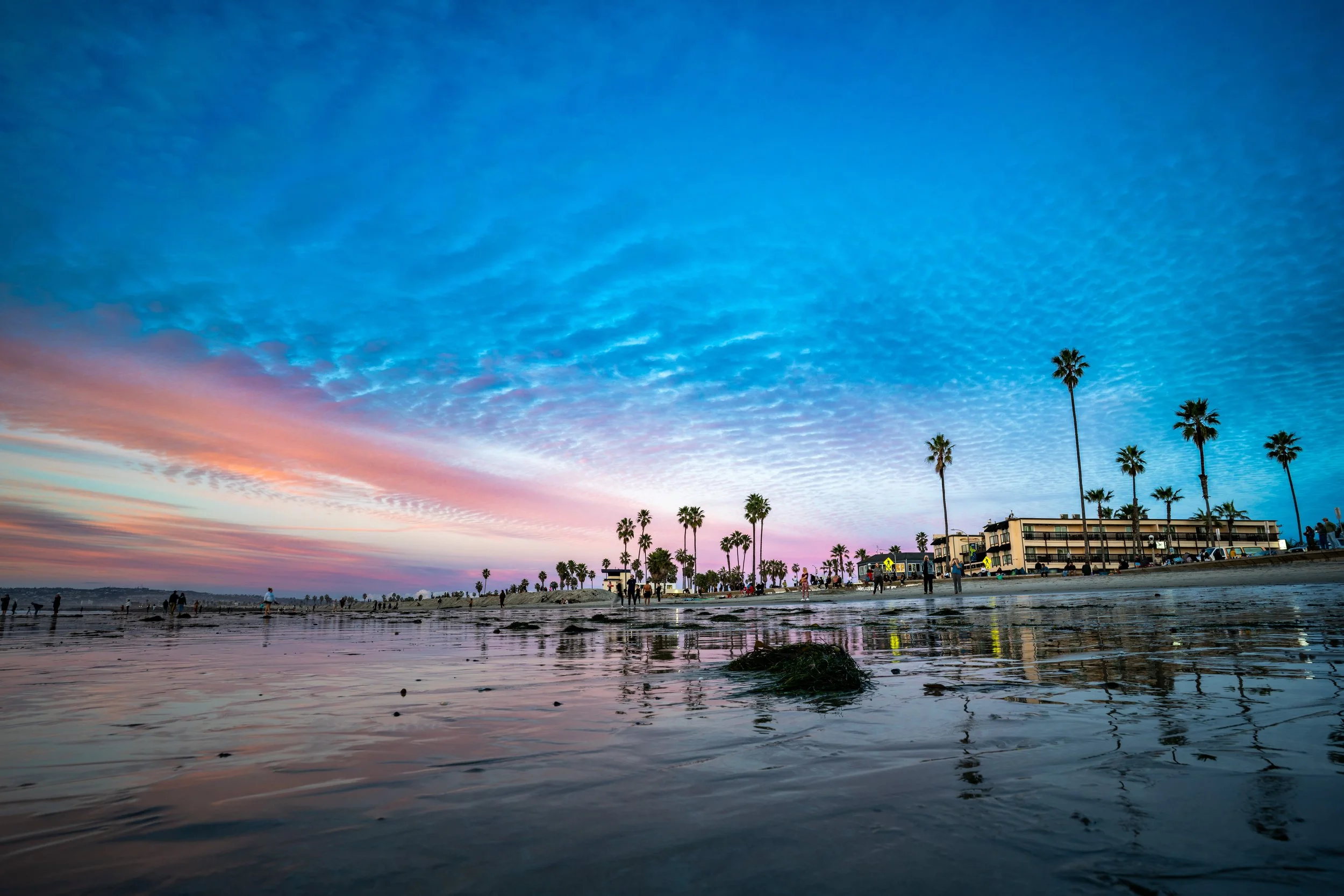 A beach at sunset with colorful sky, palm trees, and people walking along the shoreline, with buildings in the background and reflections in the water.