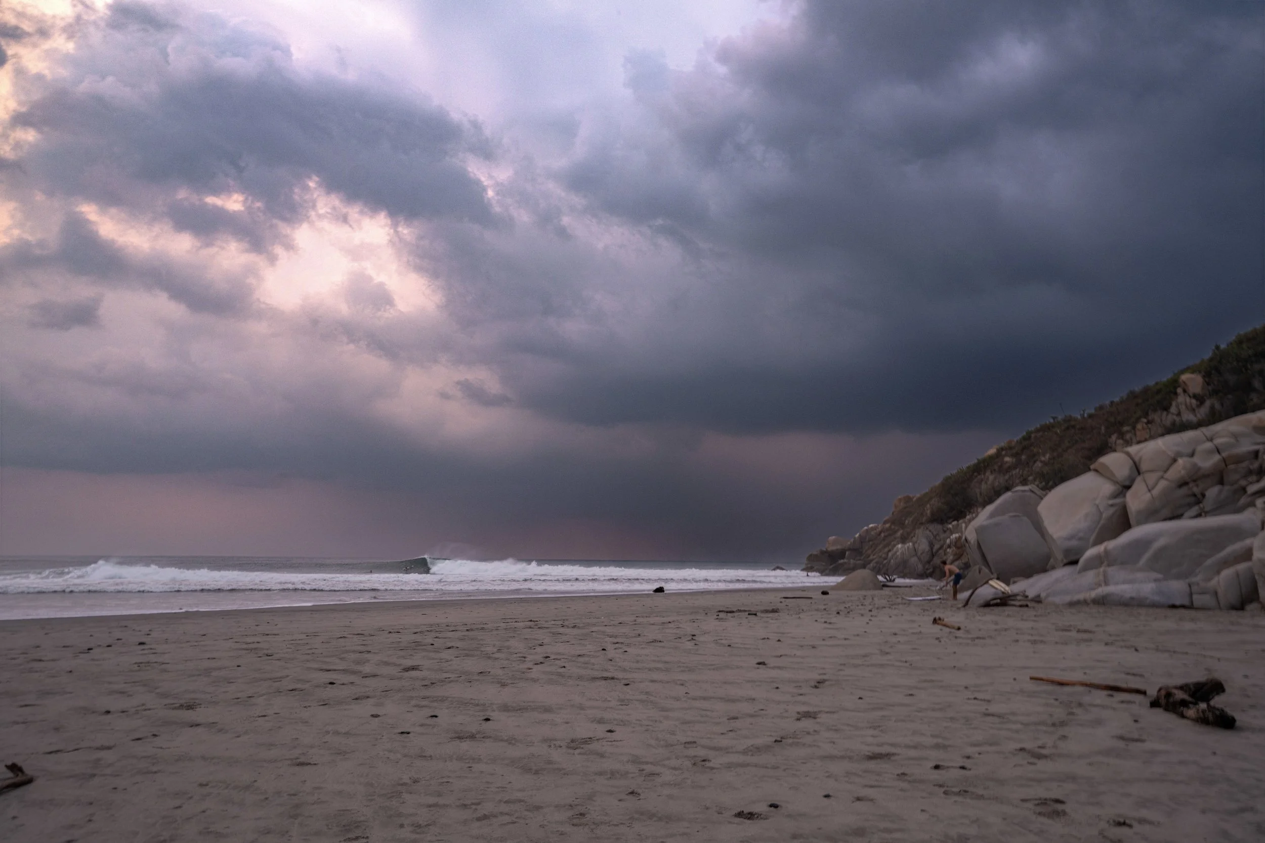 Cloudy sky over a beach with waves, rocks, and a person near the shoreline.