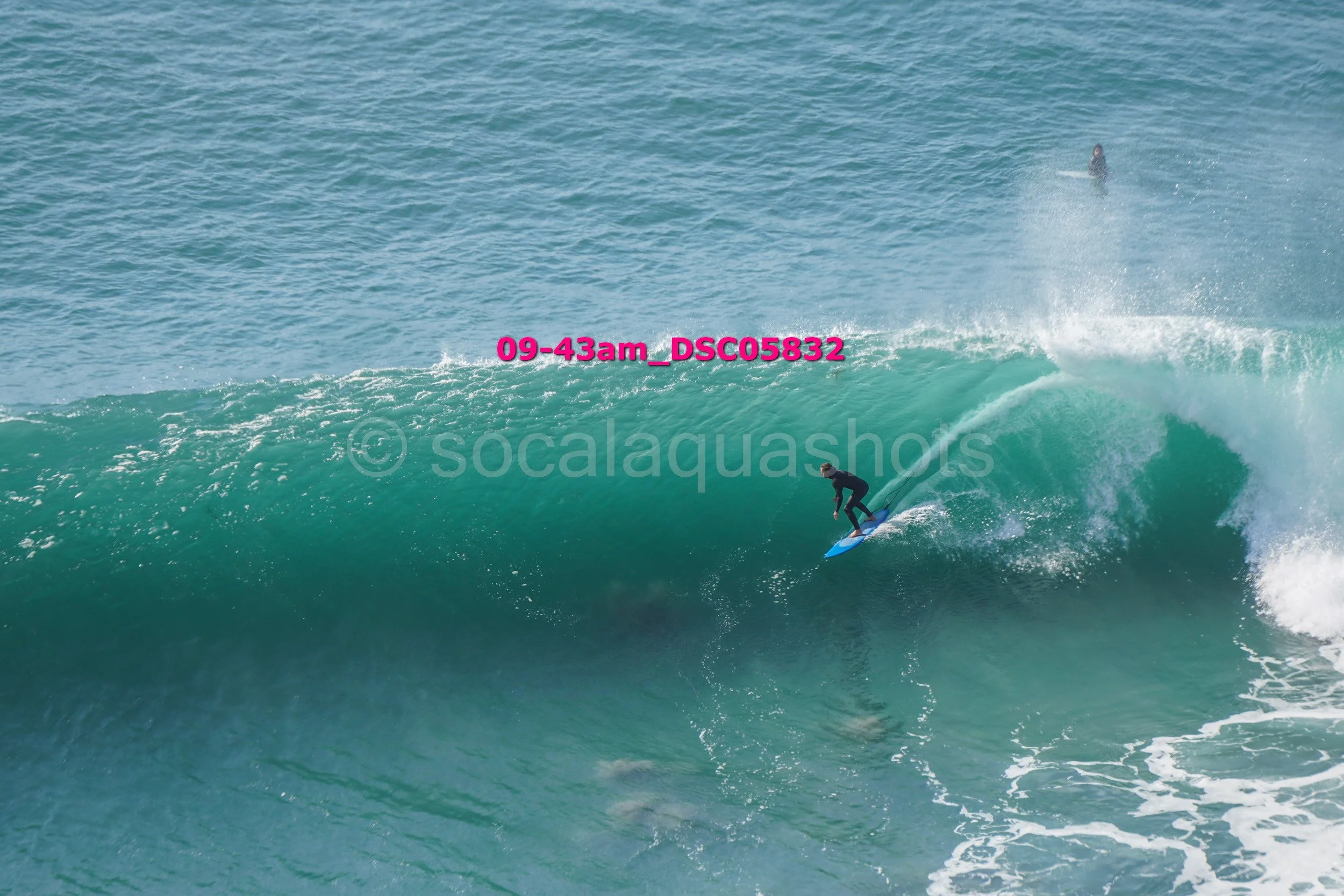A person surfing on a large wave in the ocean, with another surfer visible in the background.