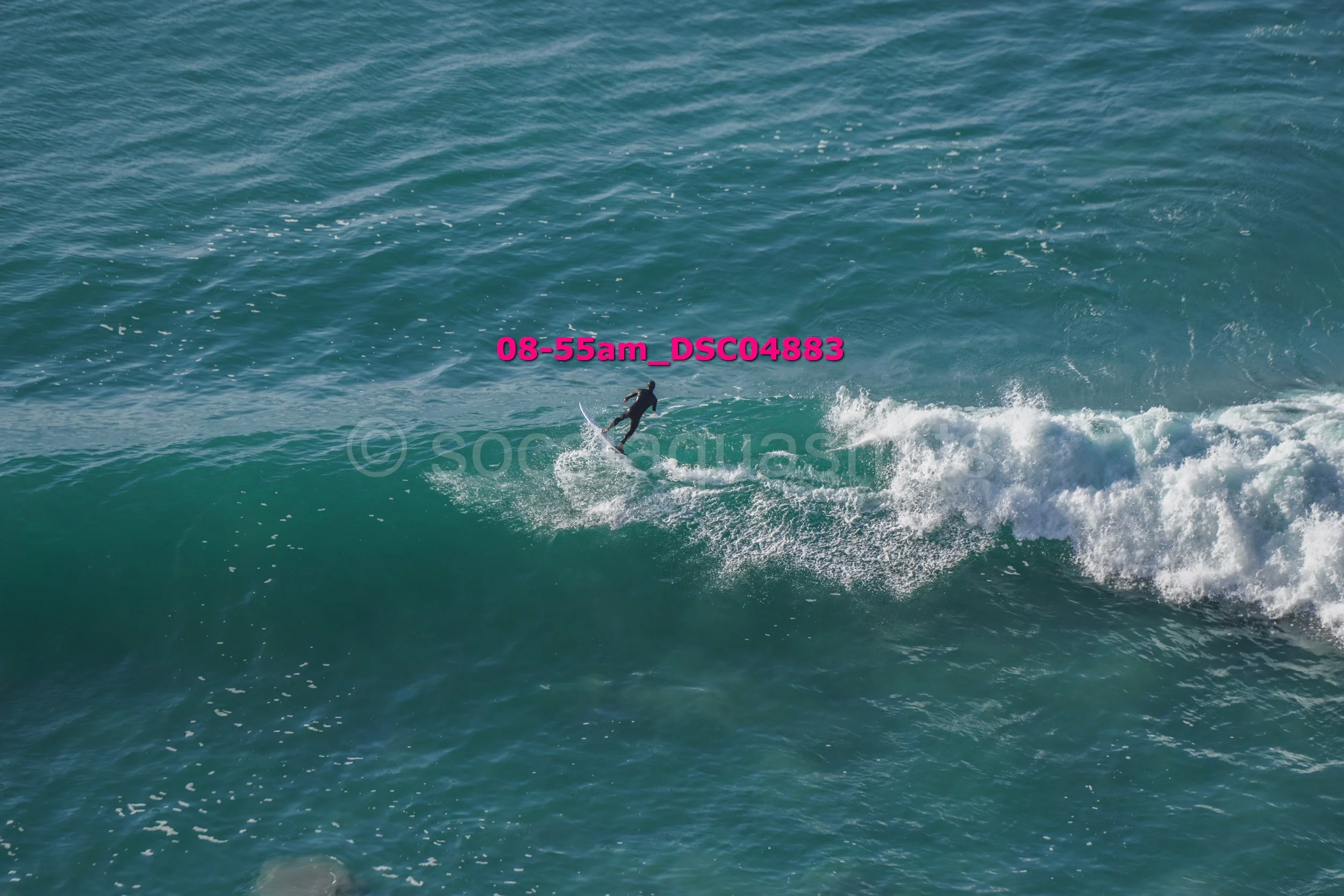 A person surfing on a wave in the ocean, wearing a wetsuit under bright daylight.