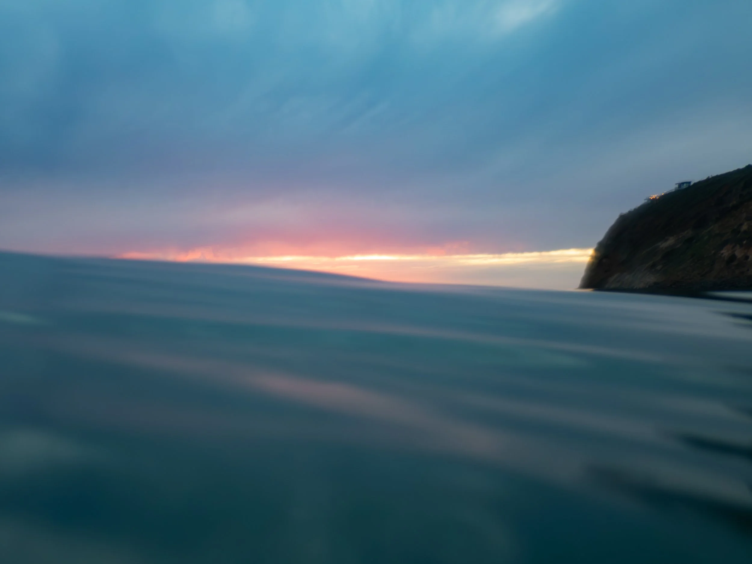 View from the water at sunset or sunrise, showing the horizon with colorful sky and a coastal cliff on the right.