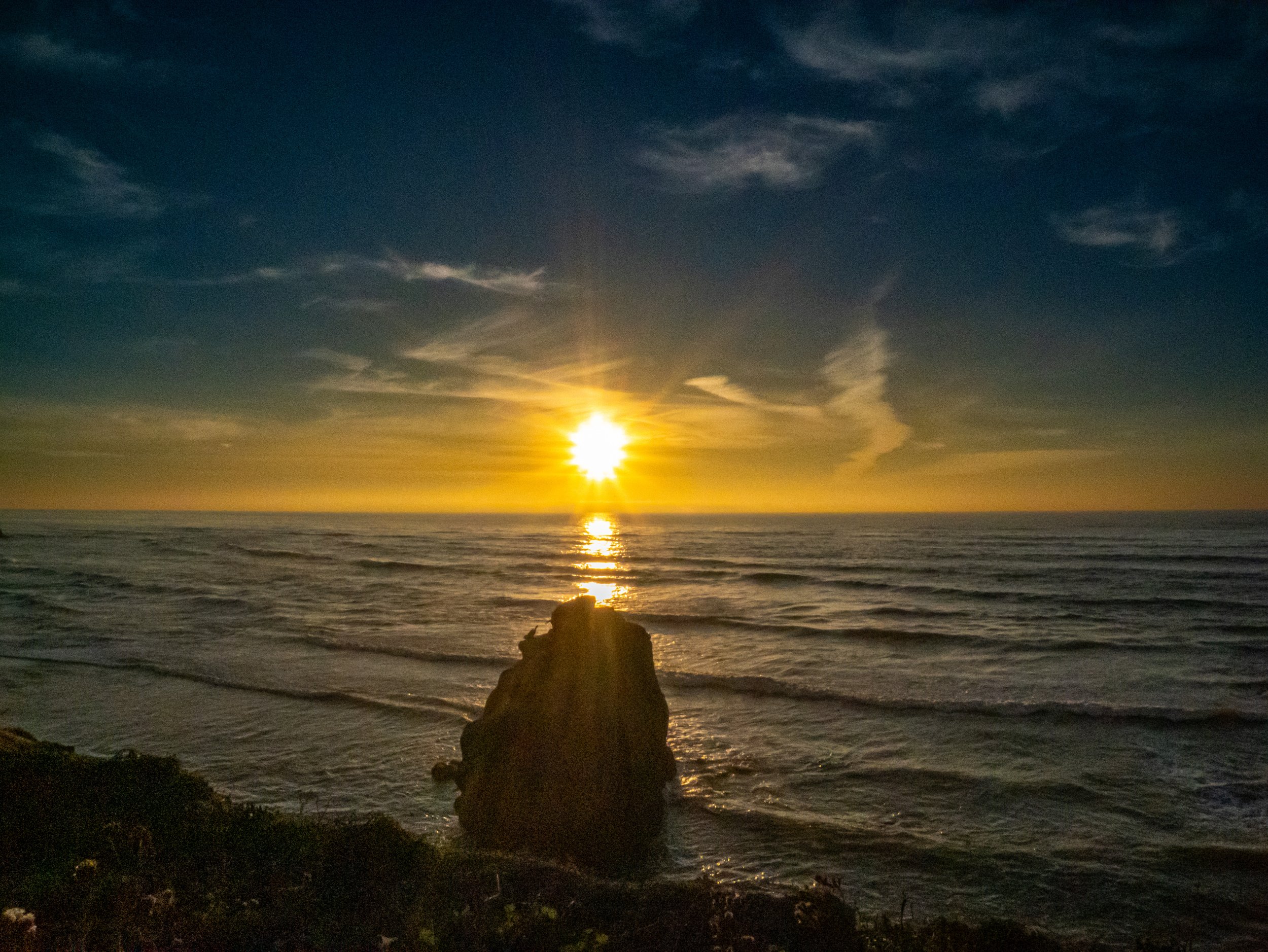 Sunset over the ocean with a large rock in the foreground and a partly cloudy sky.
