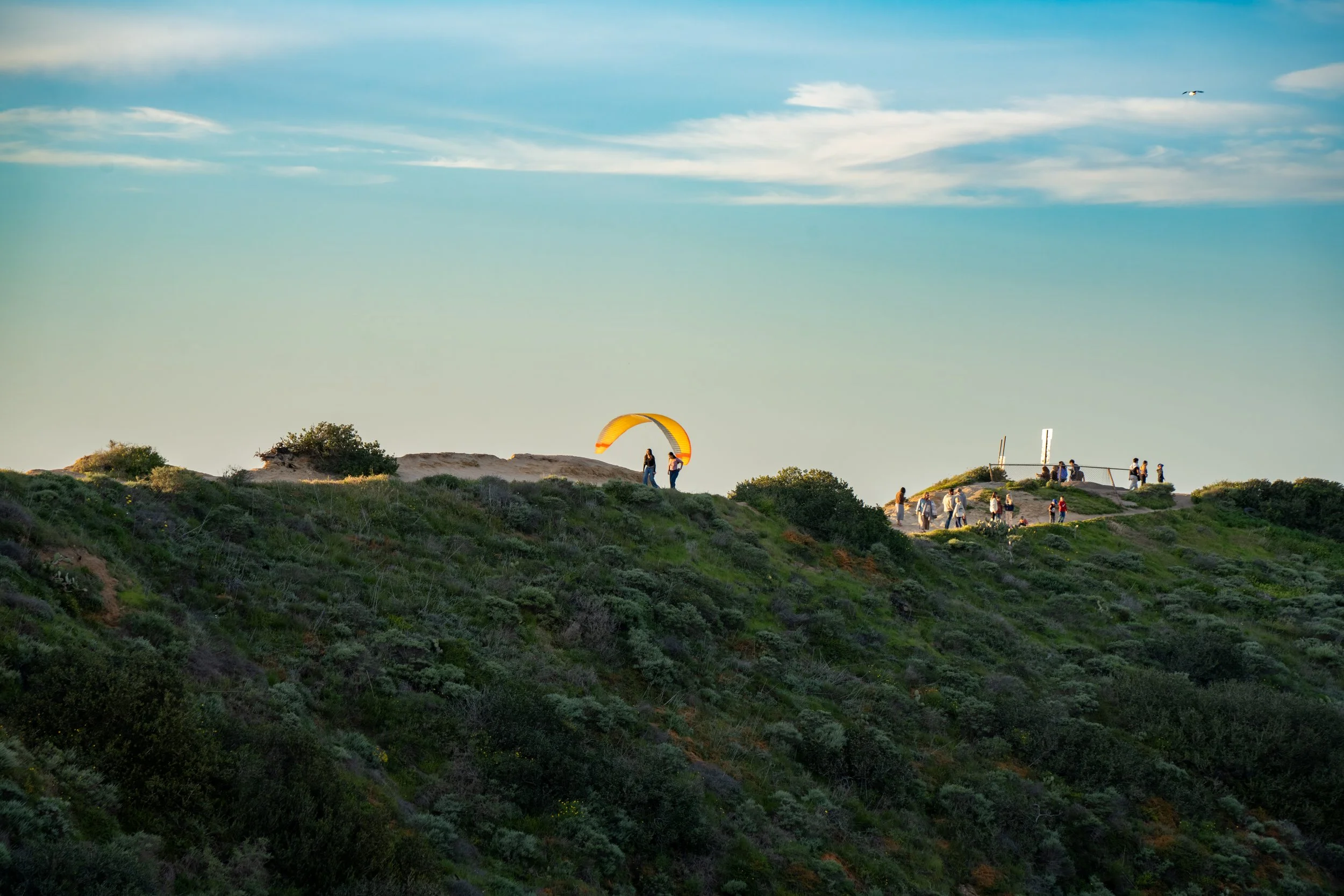 People on a green hillside with a person preparing a parachute, and others gathered near a railing in the distance, with a blue sky and clouds overhead.