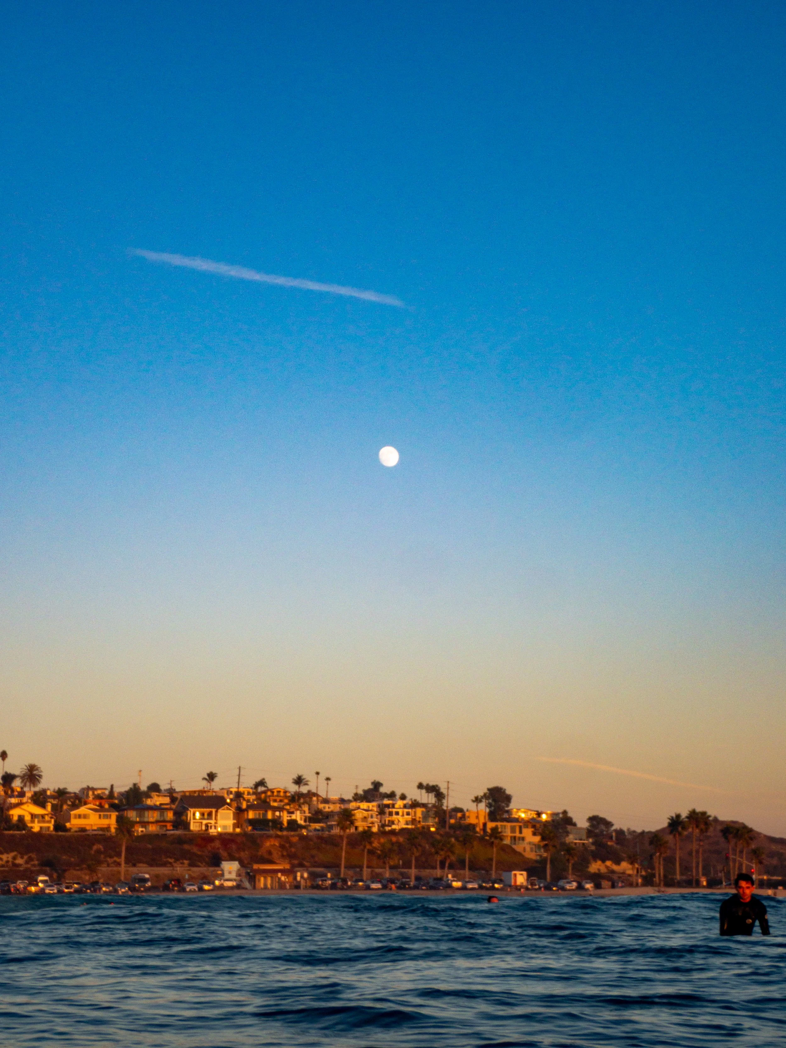 A coastal scene at sunset with the moon visible in the sky, a person in the water, and houses along the shoreline lined with palm trees.