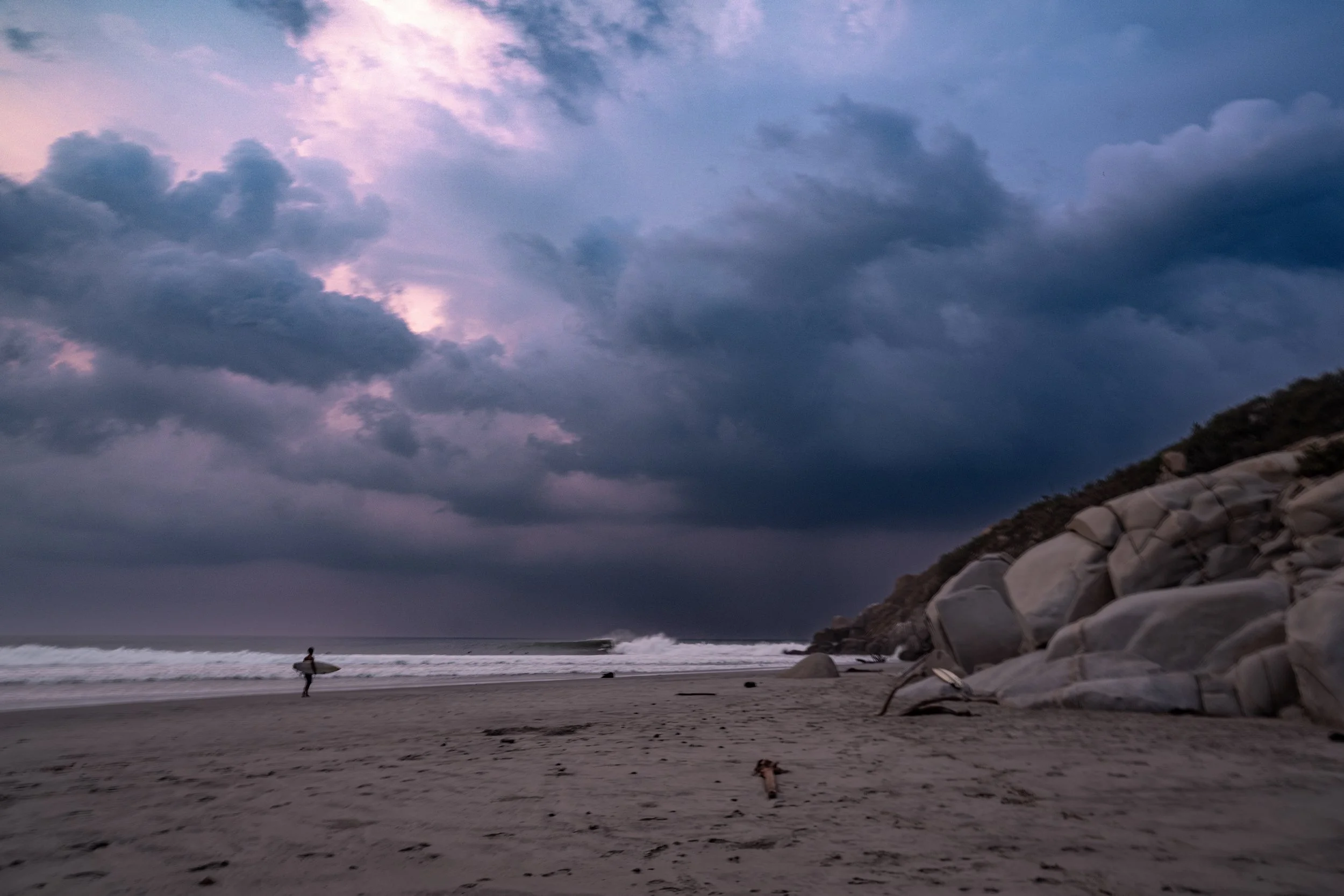 Person with a surfboard walking on the beach at dusk near rocky shoreline, cloudy sky overhead.