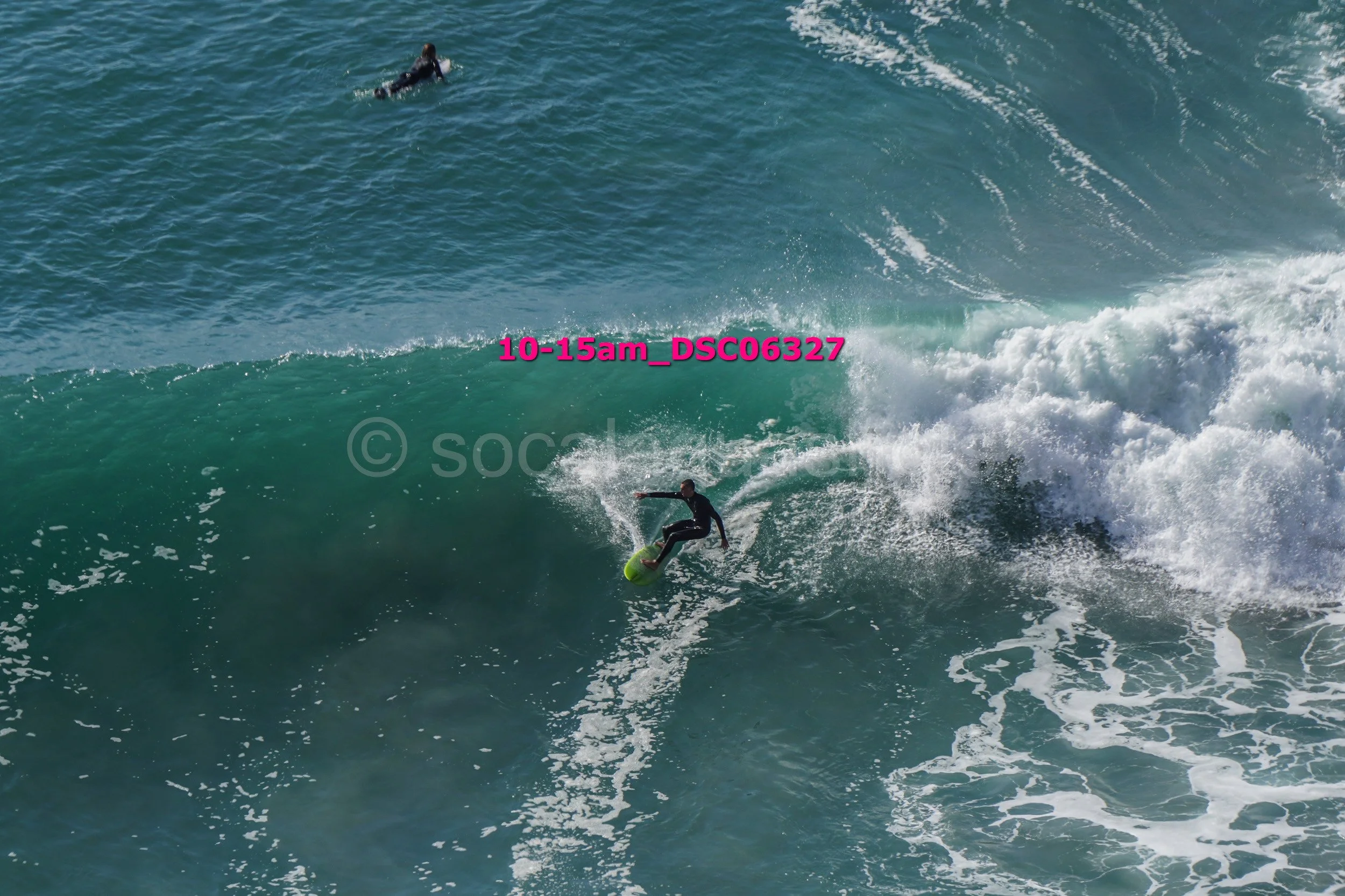 A person surfing on a large ocean wave with another surfer in the background.