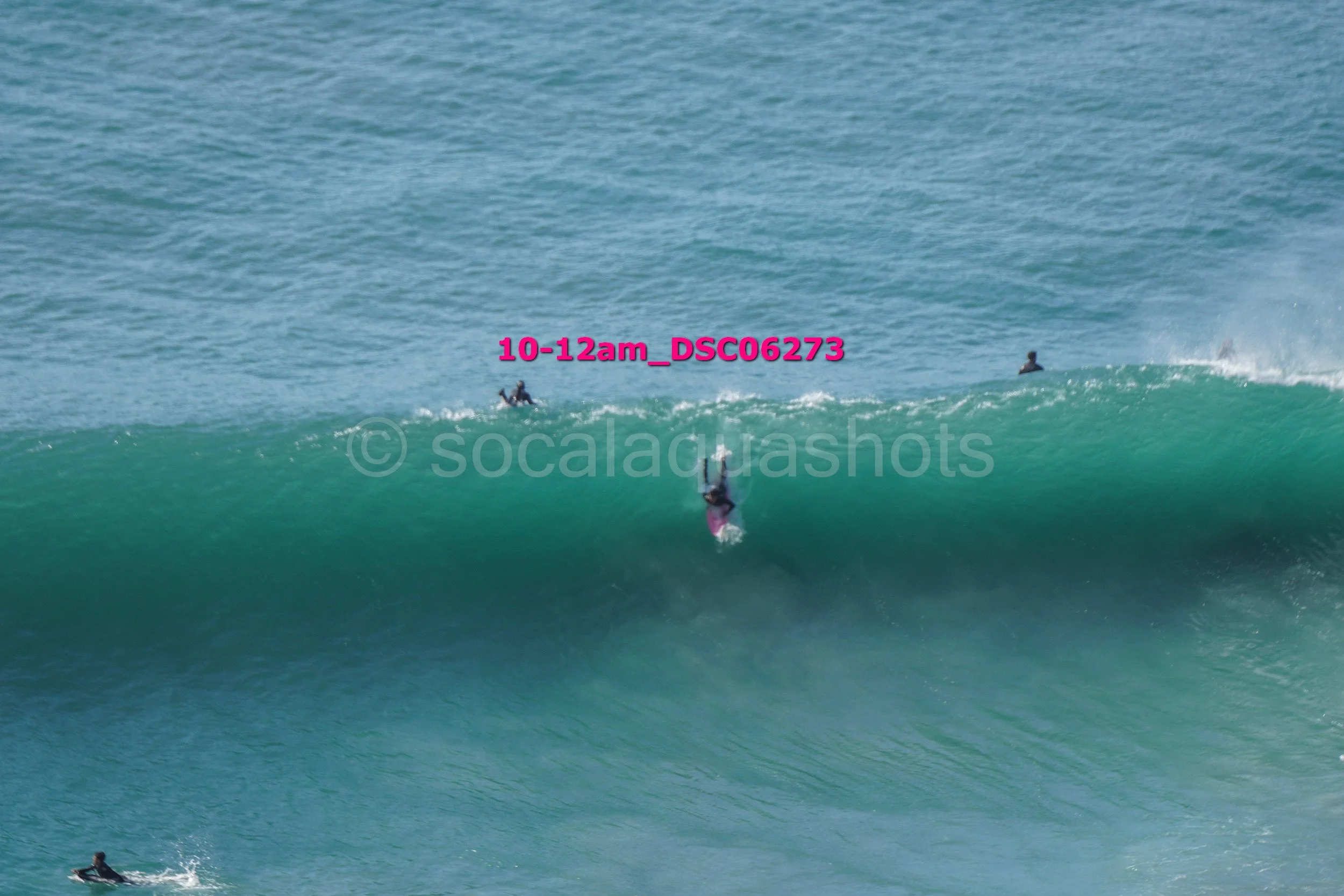 A person surfing on a large green wave in the ocean, with three other surfers visible in the background