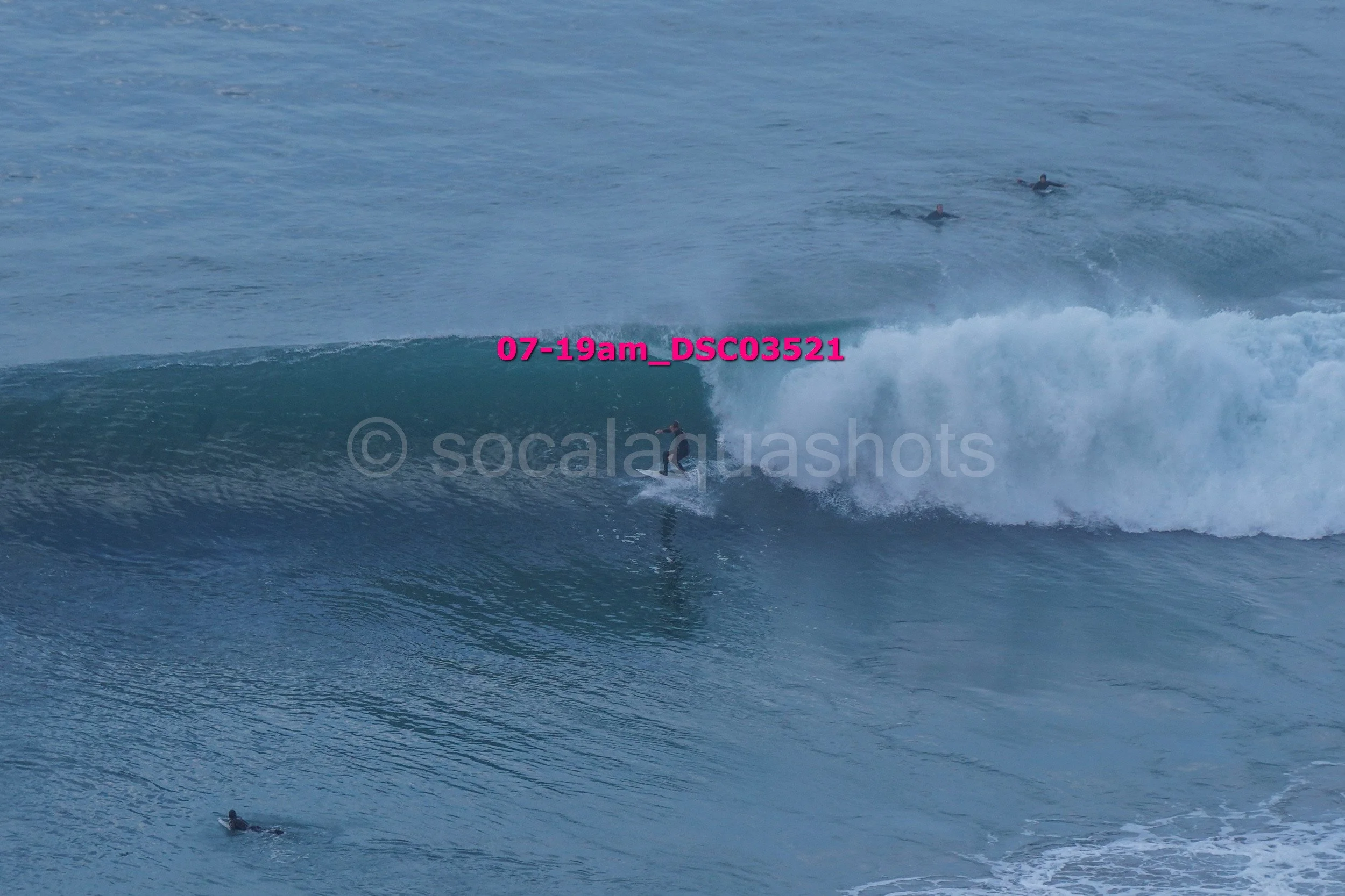 Surfer riding a wave in the ocean, with other surfers visible in the background.