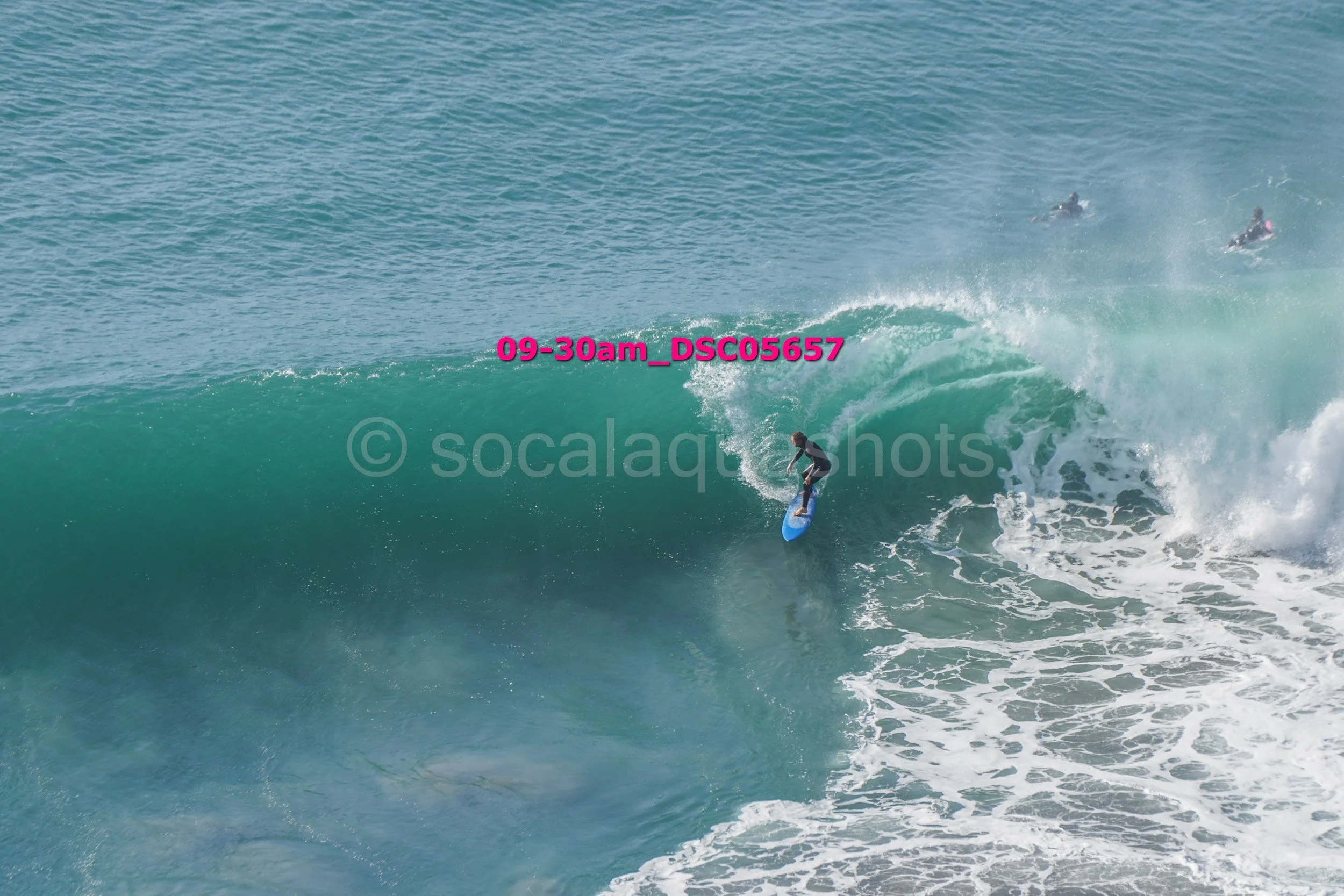 A person surfing on a large wave with two other surfers visible in the background.
