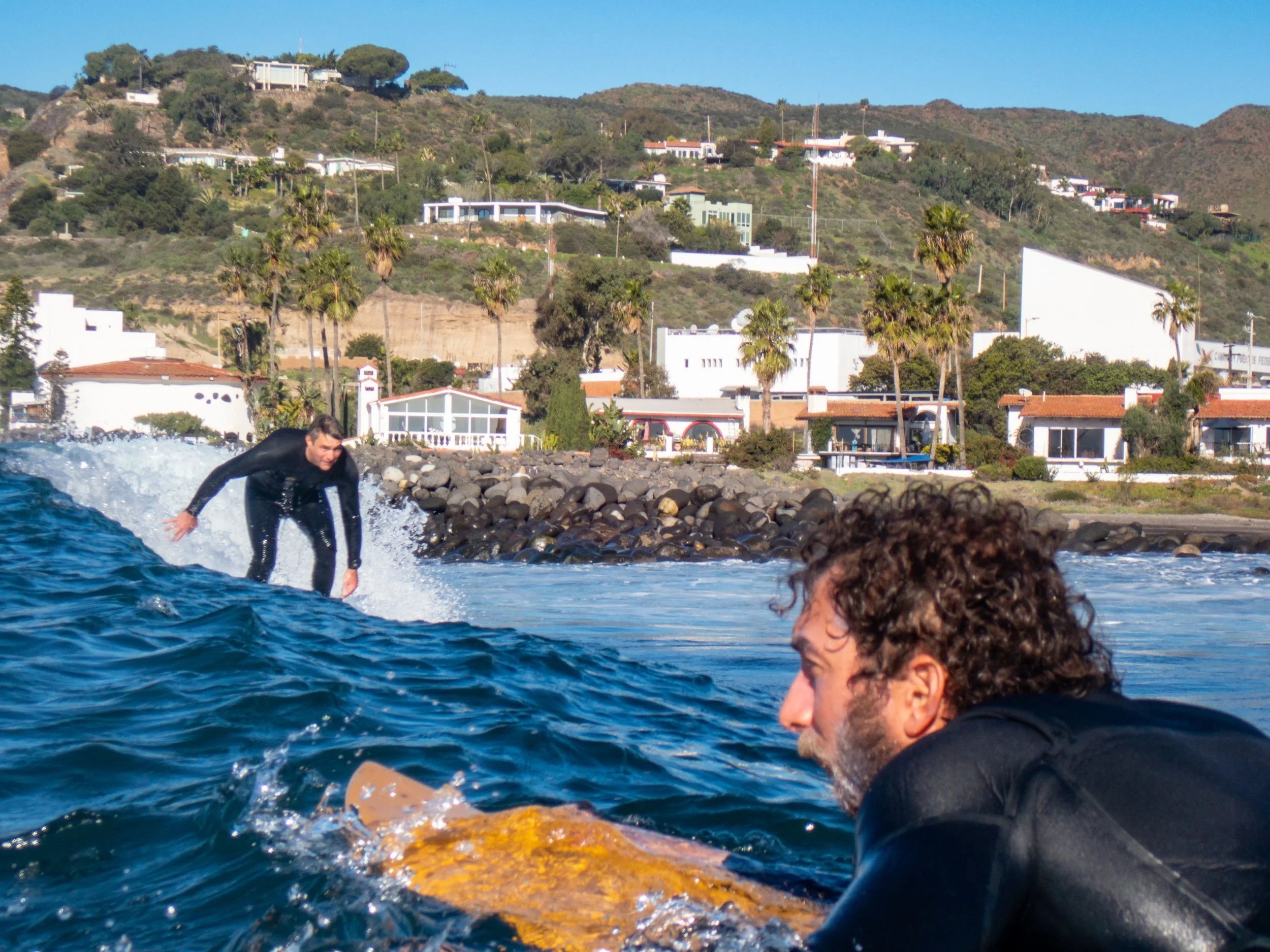 Two men in wetsuits on surfboards in the ocean near a rocky shoreline with houses and trees on a hill in the background.