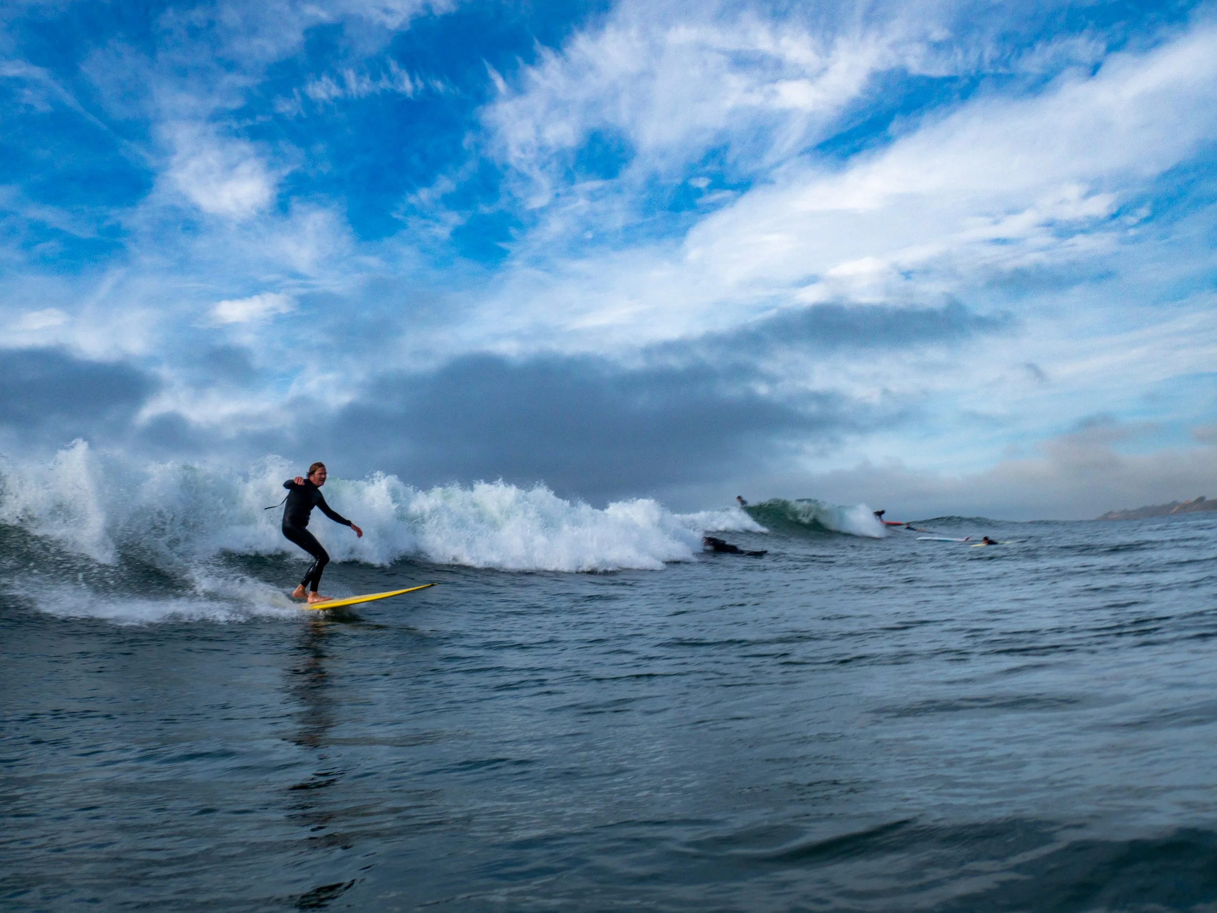 A person surfing on a wave near the coast under a cloudy sky with other surfers in the background.
