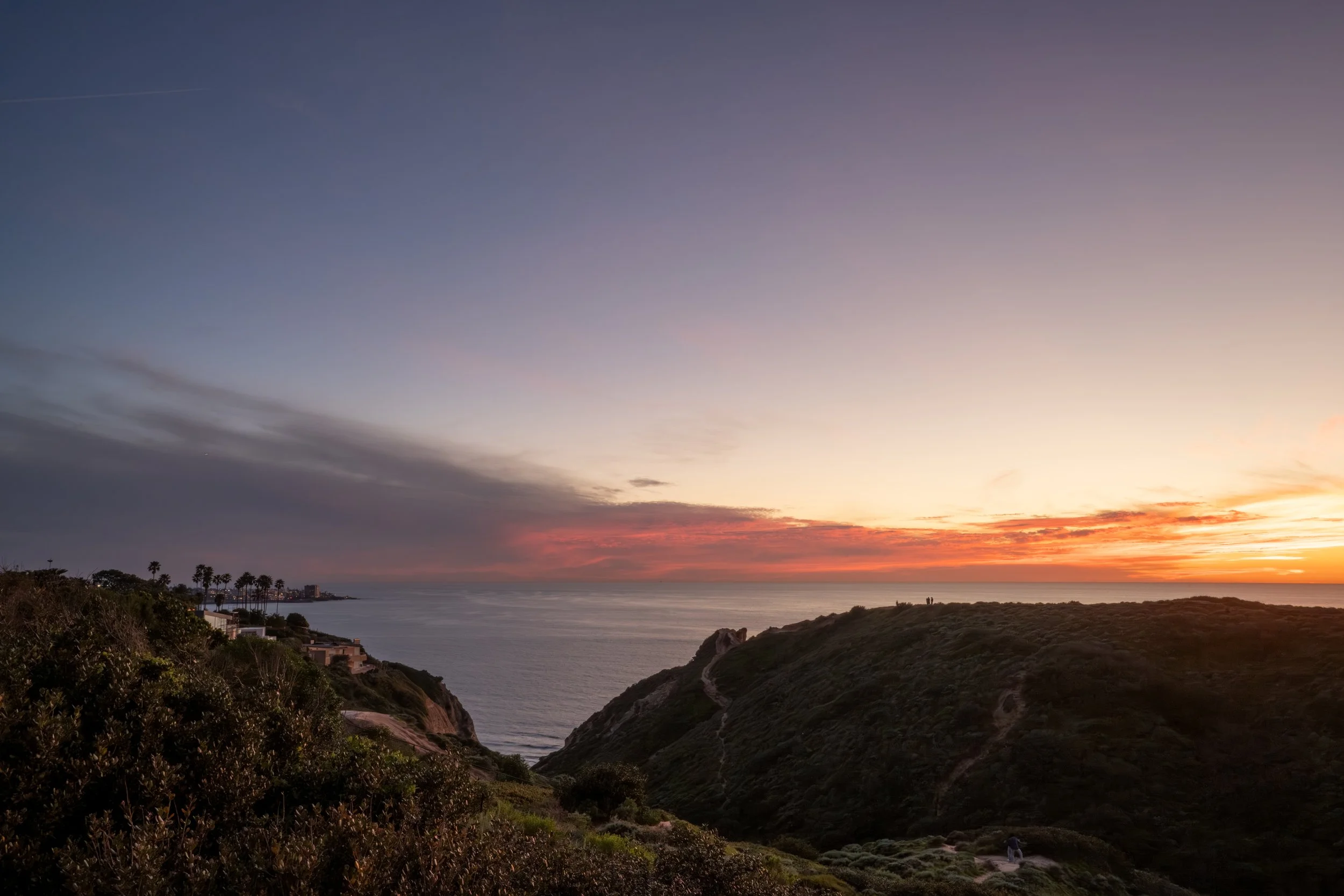 Sunset over the ocean with colorful sky and silhouetted hills and vegetation in the foreground.