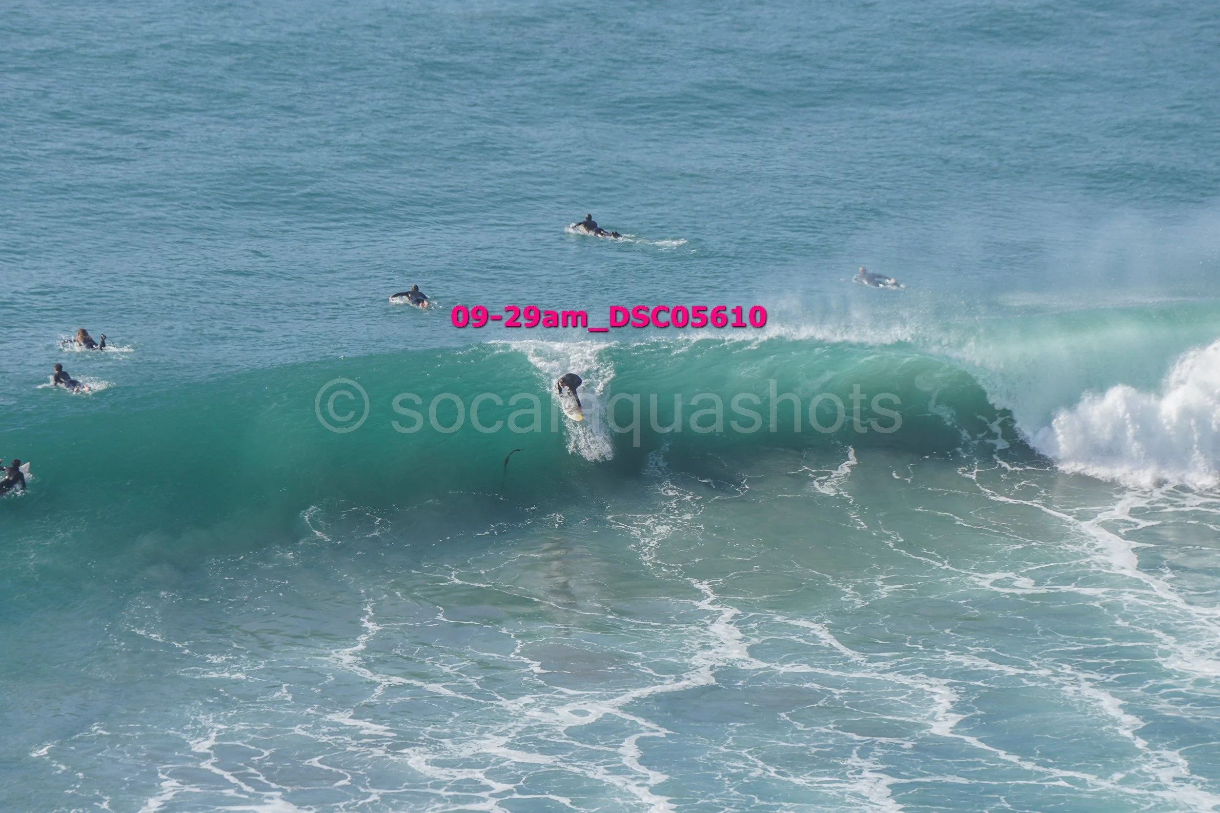 A surfer riding a large wave with several other surfers nearby in the ocean.