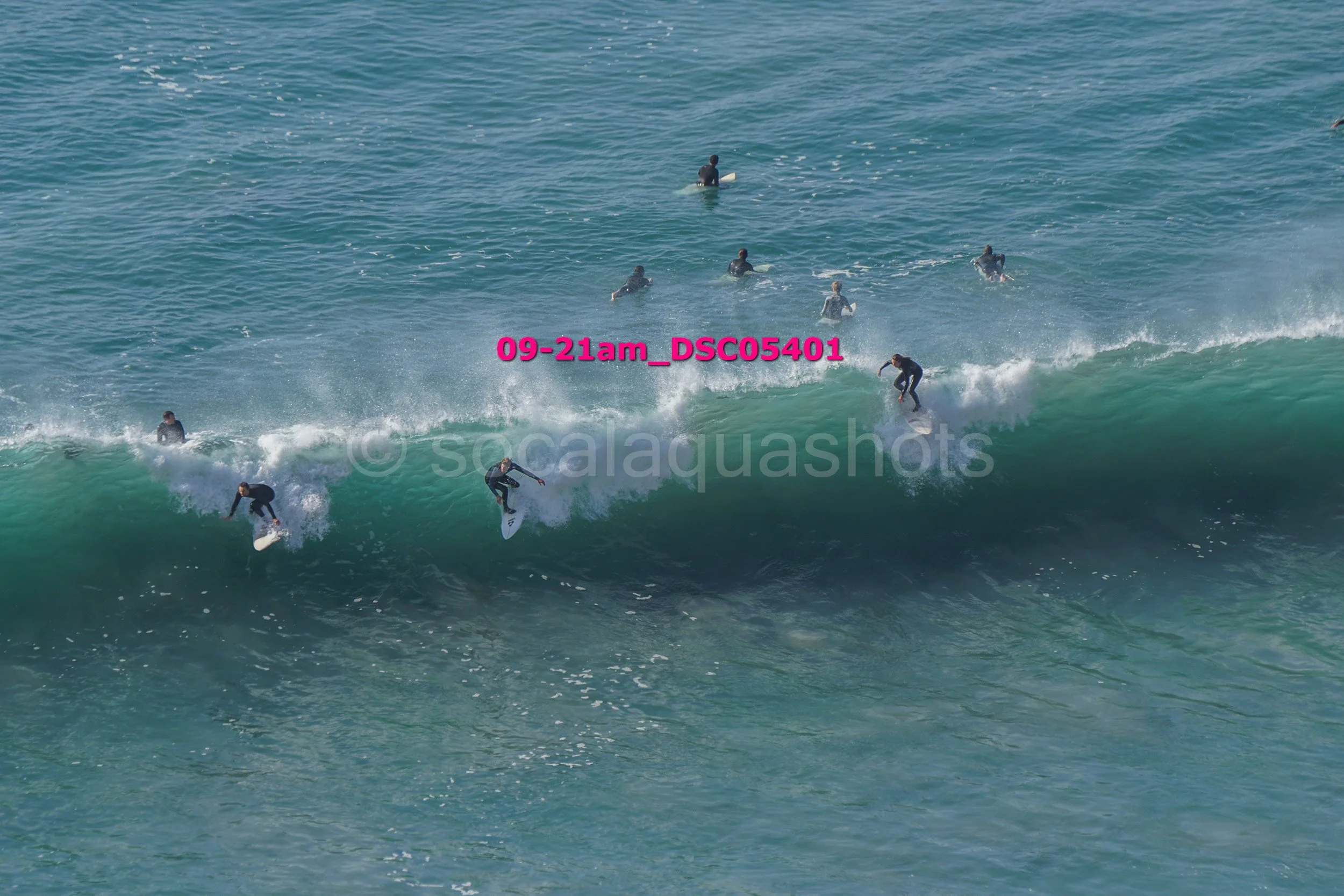 Multiple surfers riding a large wave in the ocean, with some waiting in the water and others actively surfing.