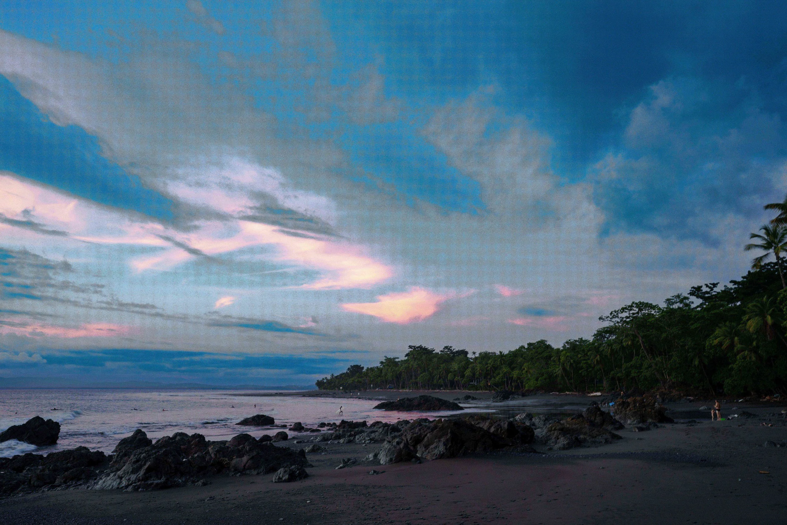 A coastal scene at sunset with dark rocks on the beach, a calm sea, and lush green trees along the shoreline. The sky has a mix of blue and pink clouds.
